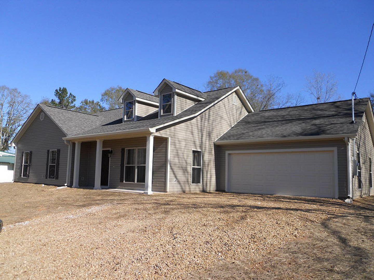Two-story house with gray siding, white-framed windows, white garage door with gray trim, brown mulch landscaping, concrete driveway, and small trees in front yard