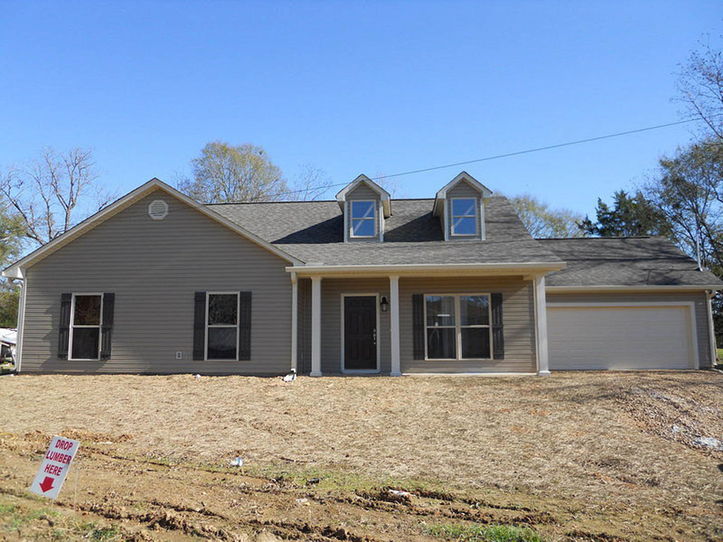 Modern two-story home with light siding, black front door framed in white, large windows, paved driveway, green lawn with a dirt patch, and a white sign with red text near the