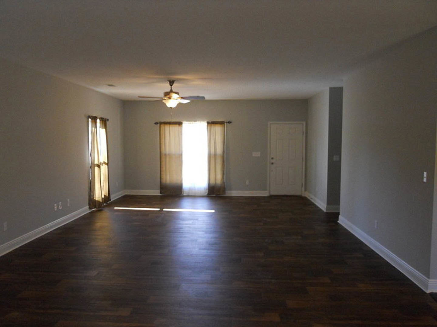 Sunlit room with dark wood flooring, white walls, ceiling fan, sheer curtain filtering light, and white door with silver doorknob