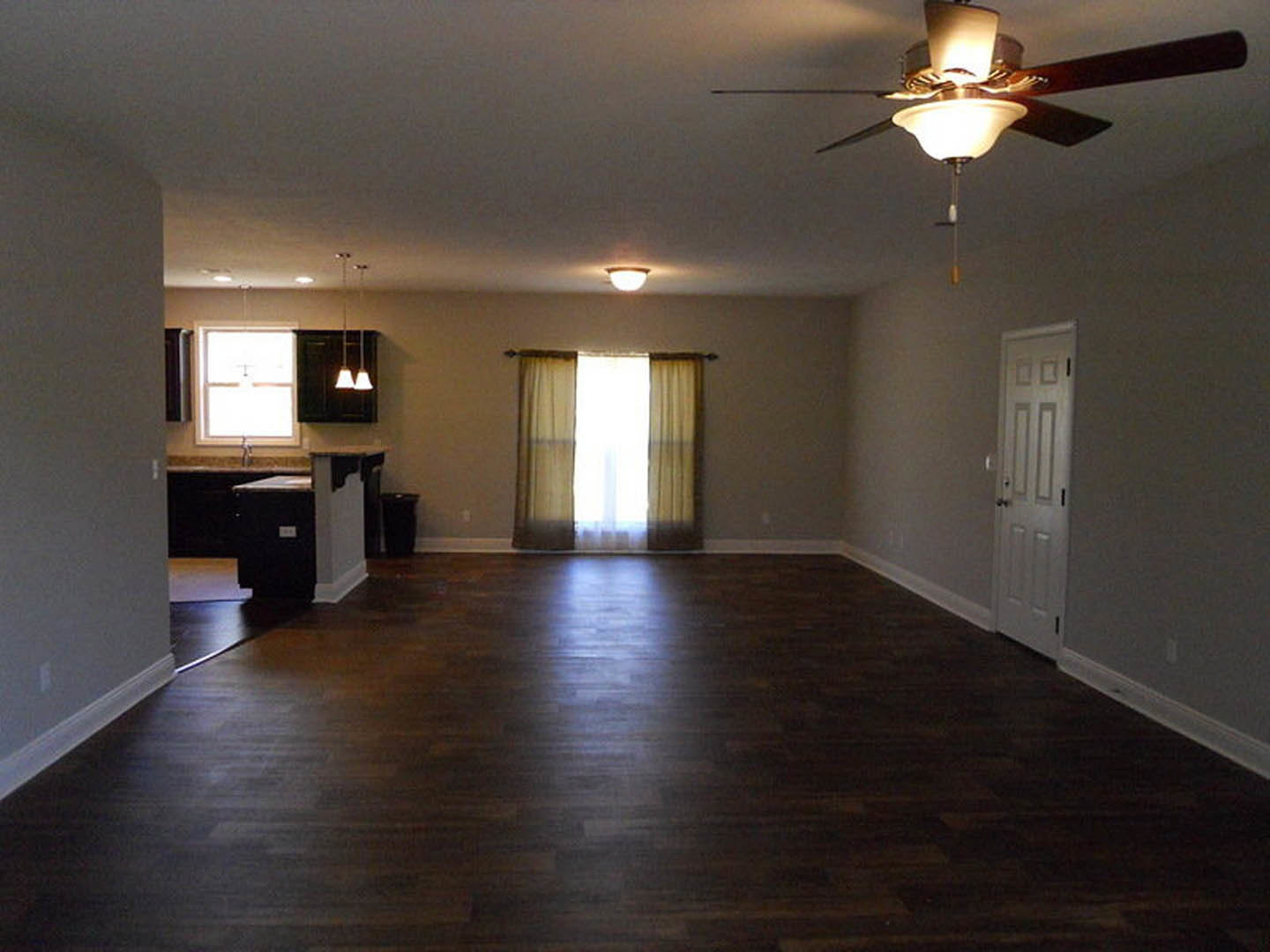 Living room with laminate flooring, ceiling fan and light fixture, open curtains on window, white door with black knobs, kitchen visible in background