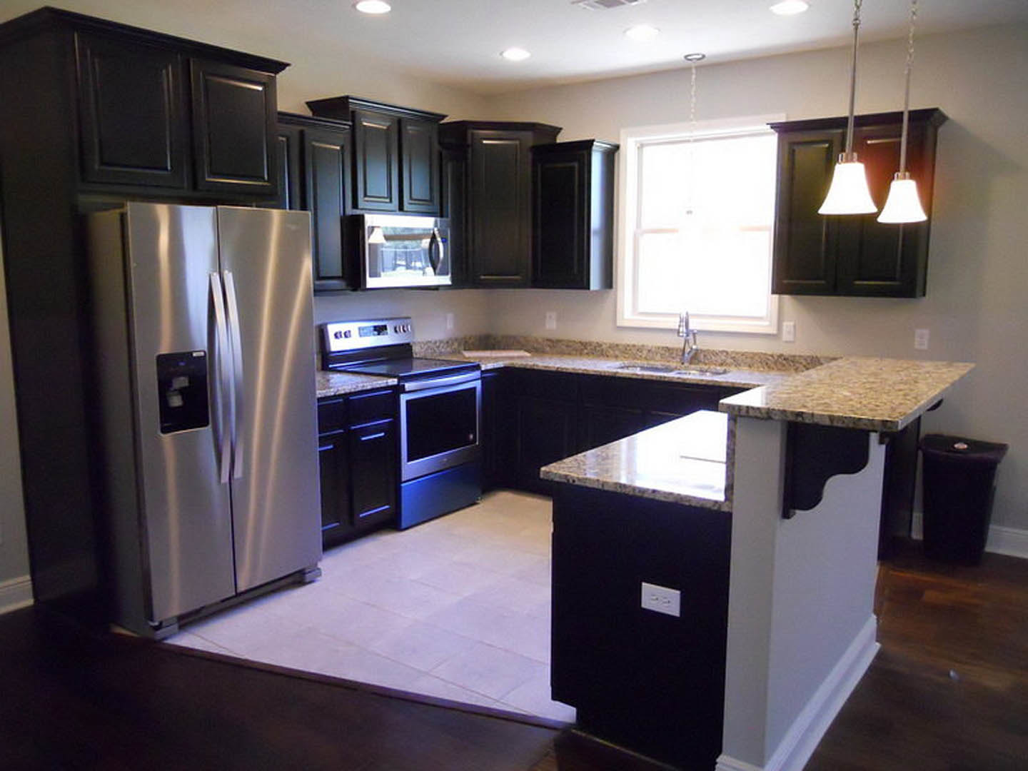 Black kitchen cabinets with granite countertops, stainless steel refrigerator, stove and oven, window with overhead light, black trash can topped with a white item, white door