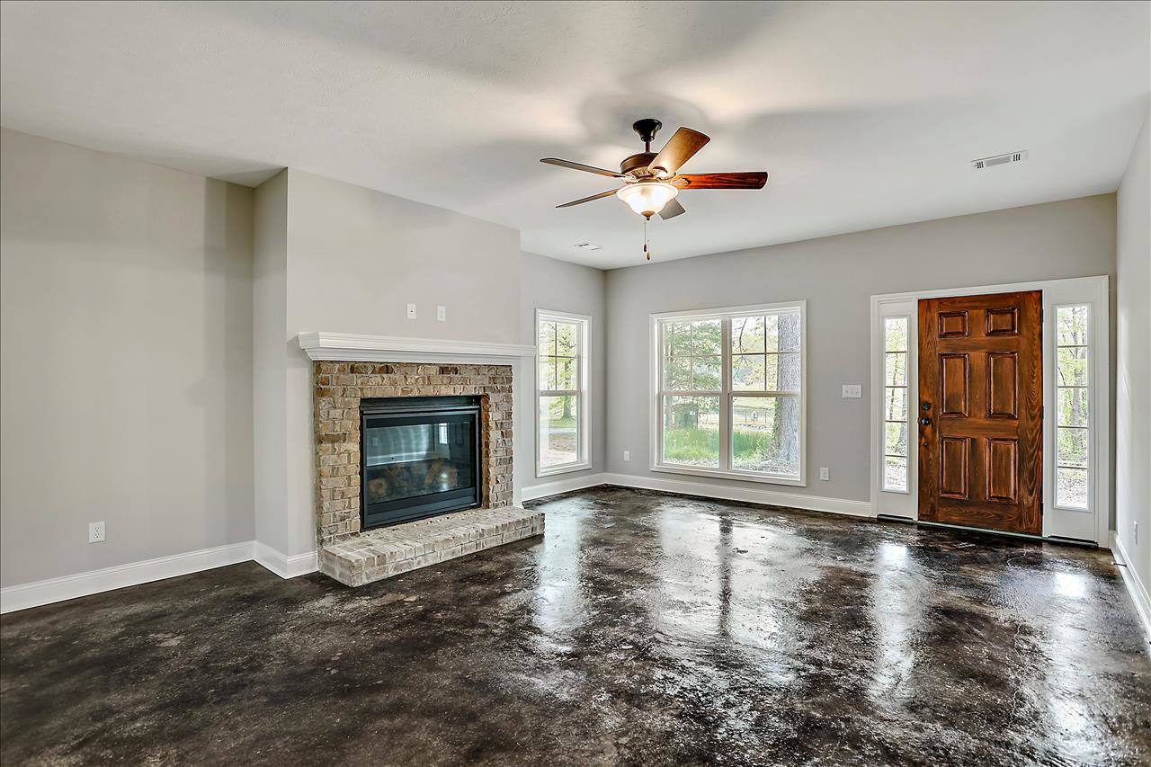 Living room with laminate flooring, ceiling fan with light, glass-door fireplace, brown door with black handle, window showing tree outside, wet floor near fireplace