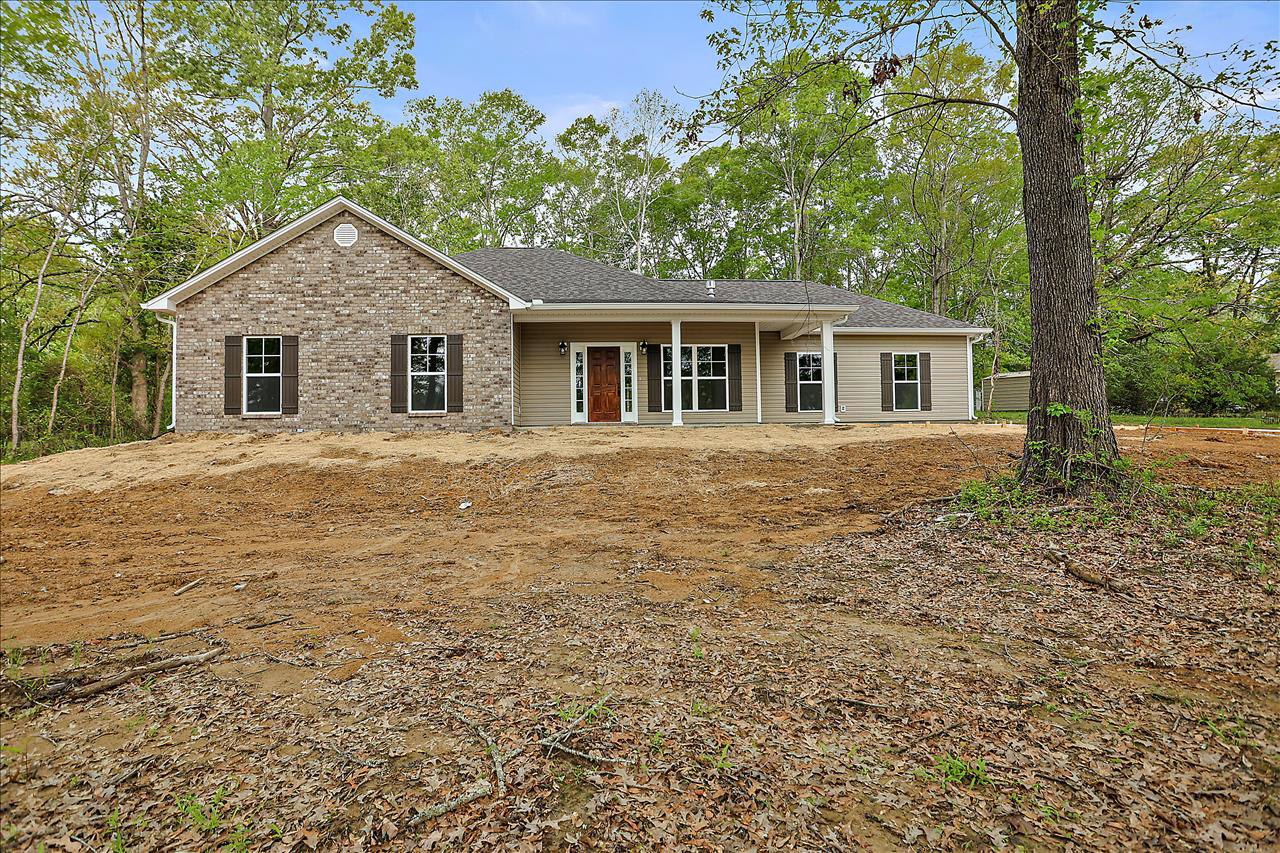 Brick house with white porch and brown door, large front yard with patchy grass and scattered leaves, mature trees and white-framed windows
