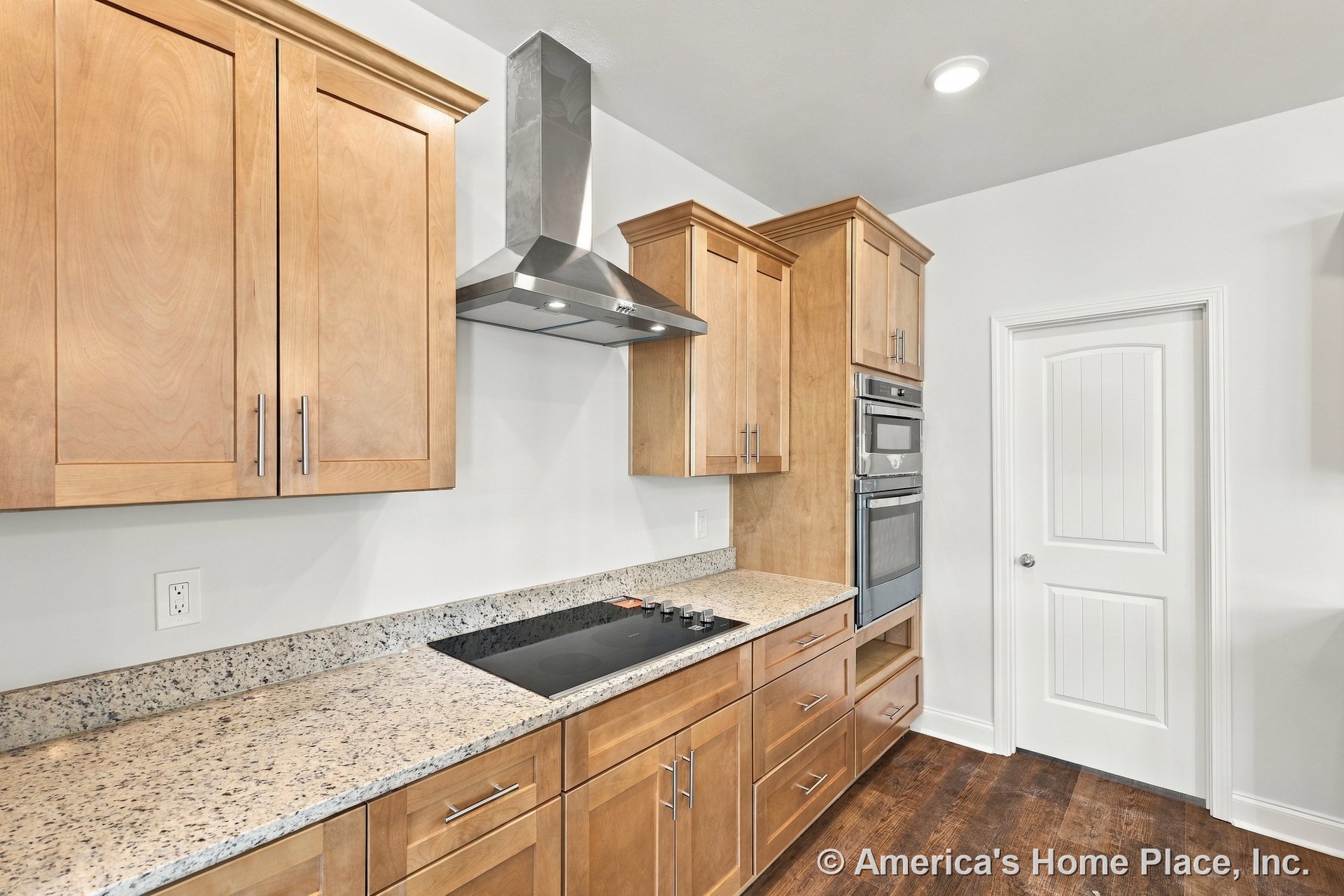 Light wood cabinetry with granite countertops, stainless steel range hood above electric cooktop, double wall ovens, white paneled door, wood flooring, recessed lighting, and
