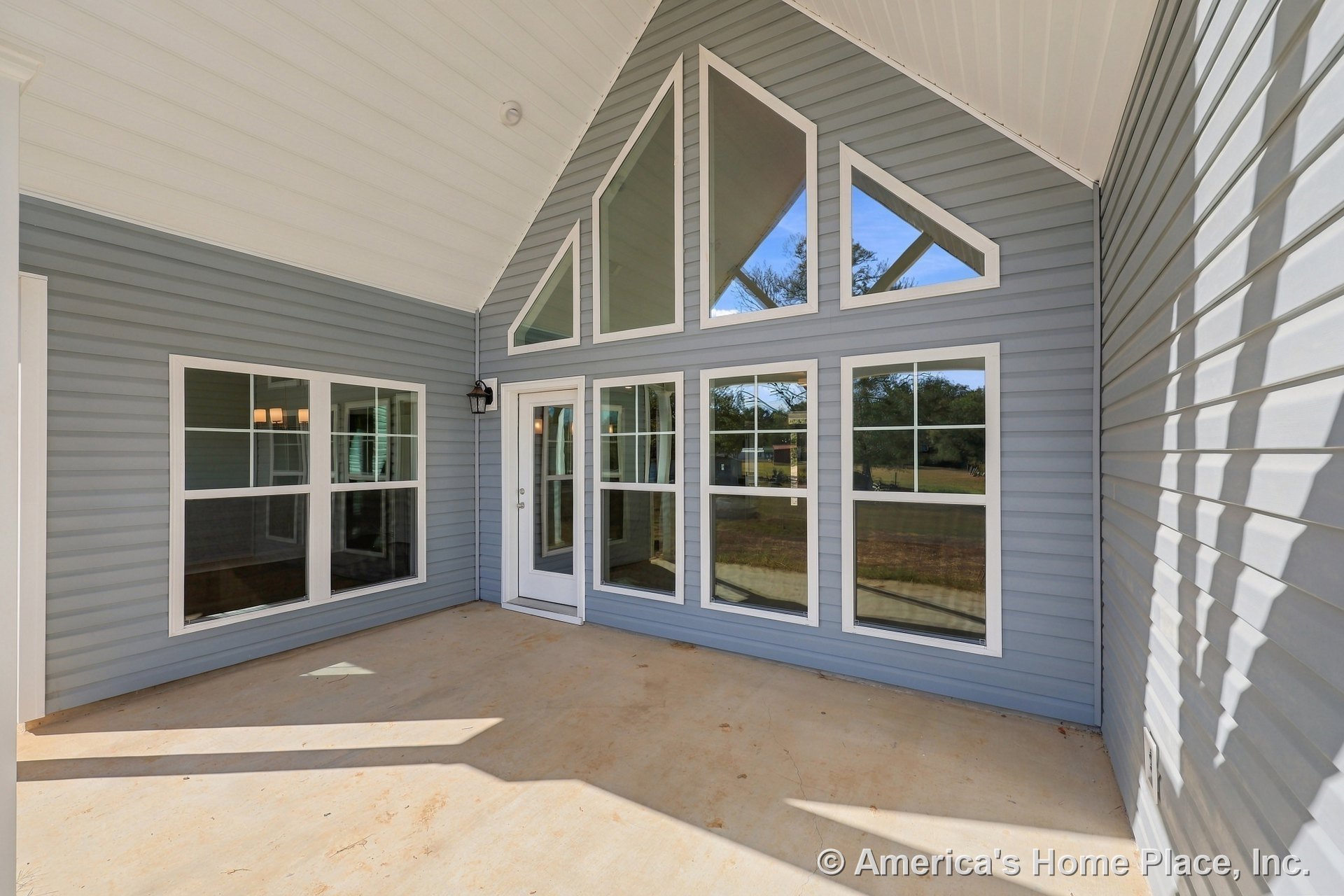 Covered porch with vaulted white ceiling, gray horizontal siding, triangular transom windows, multiple grid-style geometric windows, single glass door, concrete flooring, black