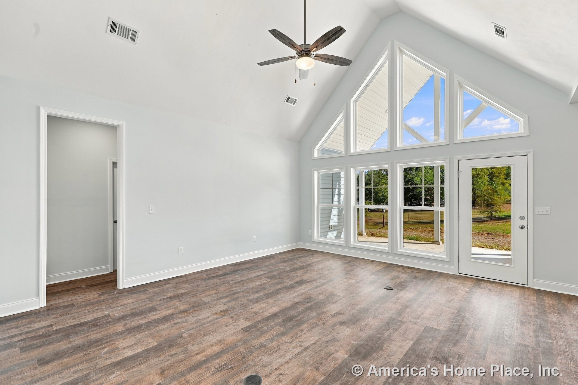 Vaulted ceiling living room with large triangular windows, wood-look flooring, white trim and baseboards, ceiling fan with light, glass exterior door, and transom windows allowing