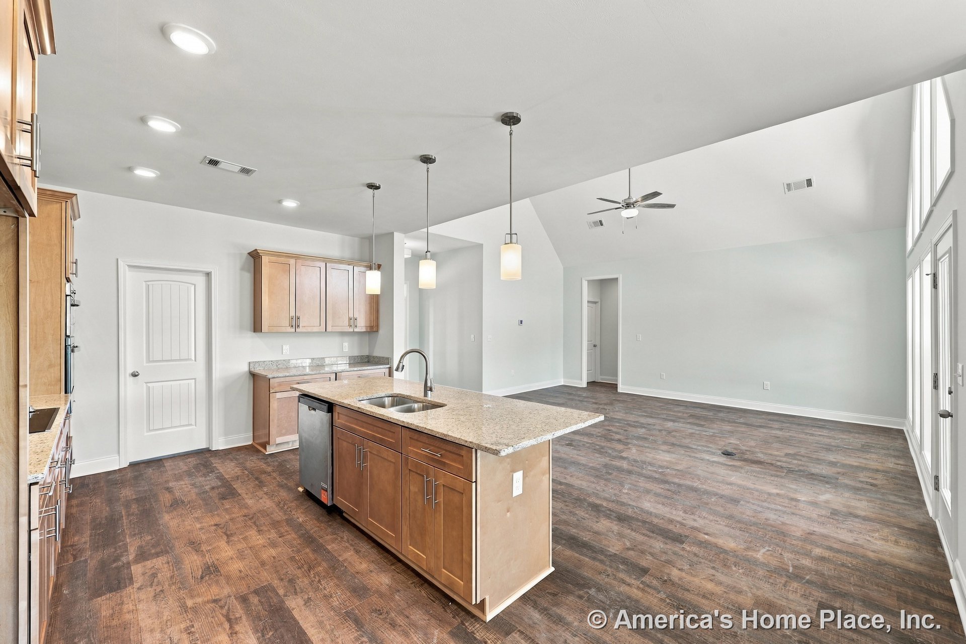 Wood kitchen cabinetry and granite island countertop with pendant lights, open to a living area featuring vaulted ceilings, wide plank wood flooring, large windows, and glass doors