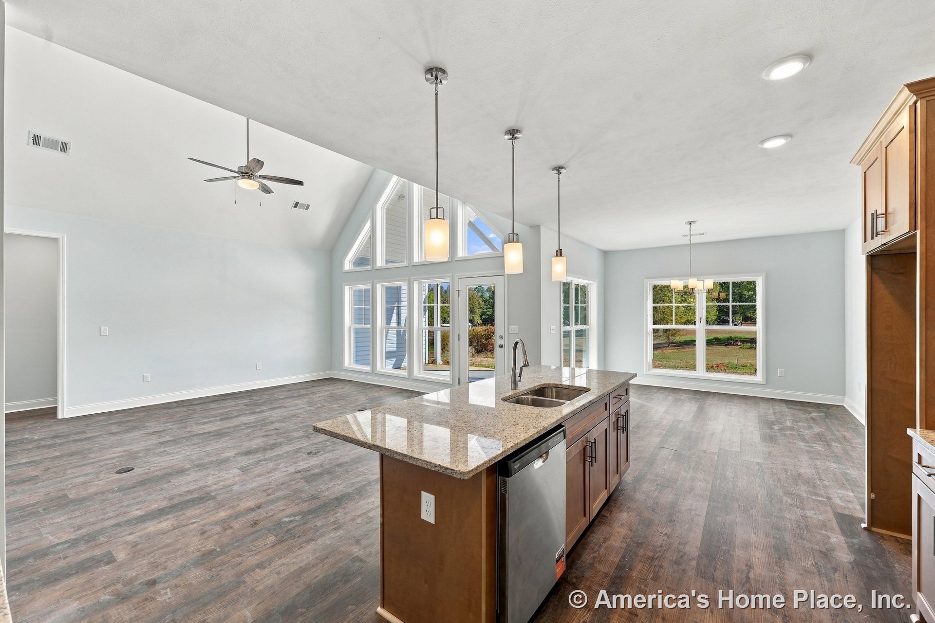 Granite kitchen island with double sink and dishwasher, wood cabinetry, pendant and recessed lighting, open layout connecting to living area with vaulted ceiling fan, wall of large