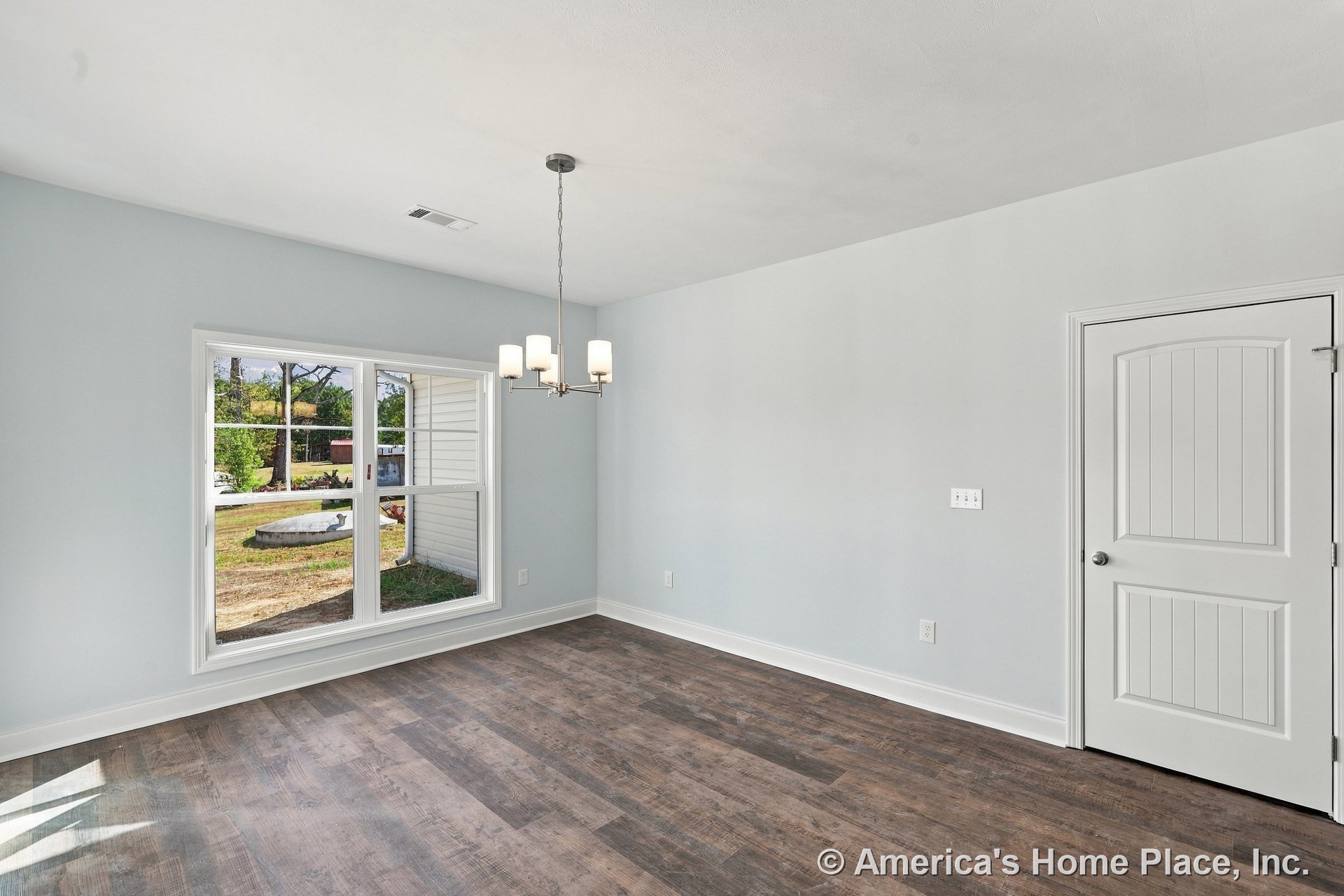 Dining area with large double window, wood-look plank flooring, modern ceiling light fixture, white paneled door, light gray painted walls, and white baseboards.