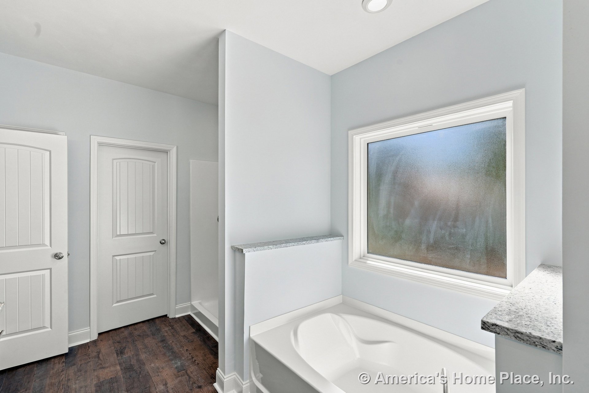 Built-in soaking tub set beneath a frosted window with granite ledges, adjacent walk-in shower, white paneled doors, dark wood flooring, recessed lighting, and white walls.
