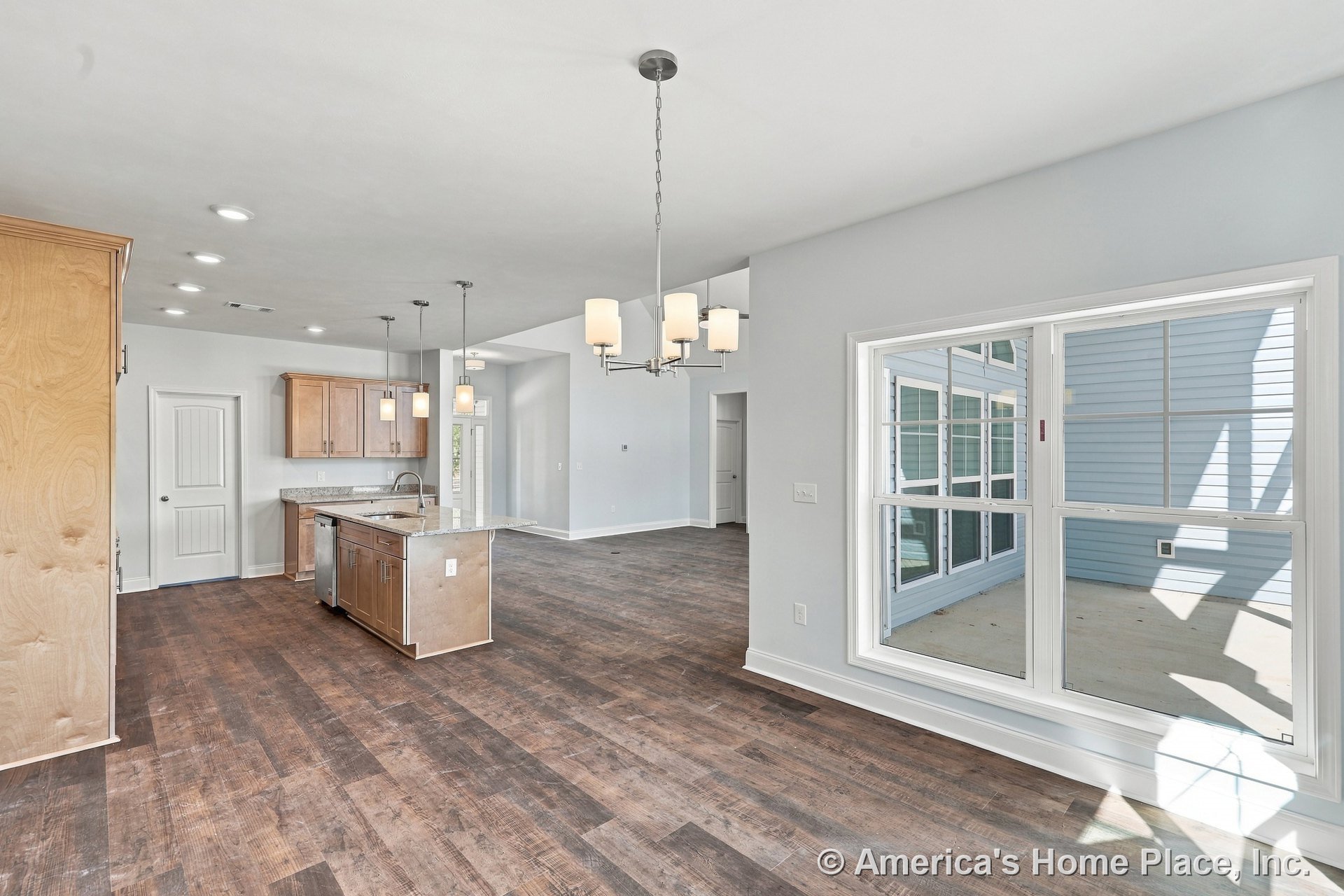 Open kitchen and dining area with wood cabinetry, granite countertops, wide plank wood flooring, kitchen island with sink, double window, pendant and chandelier lighting.