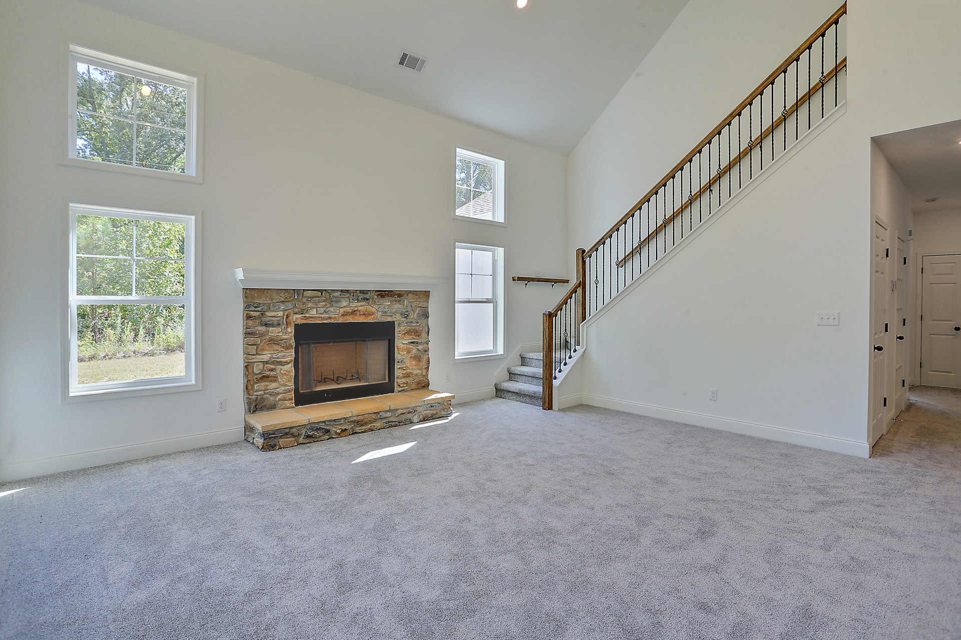 Living room with light wood flooring, white plaster walls, black-framed fireplace with screen, staircase with wood treads, large window showing trees and roof outside