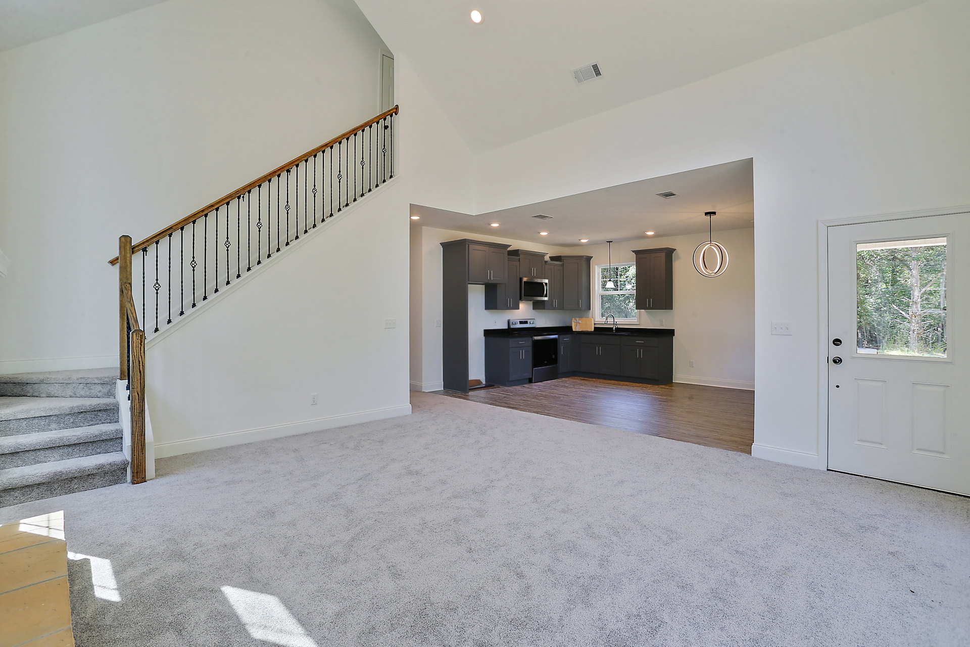 Open concept room featuring a kitchen with laminate flooring, white cabinetry, and ceiling light; adjacent staircase with metal railing; white door with window; large window