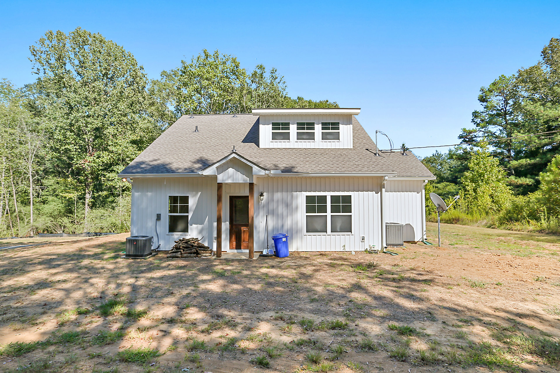 Two-story house with beige siding, white trim, and multiple square windows, surrounded by green lawn, mature trees, and a blue trash can near the side.