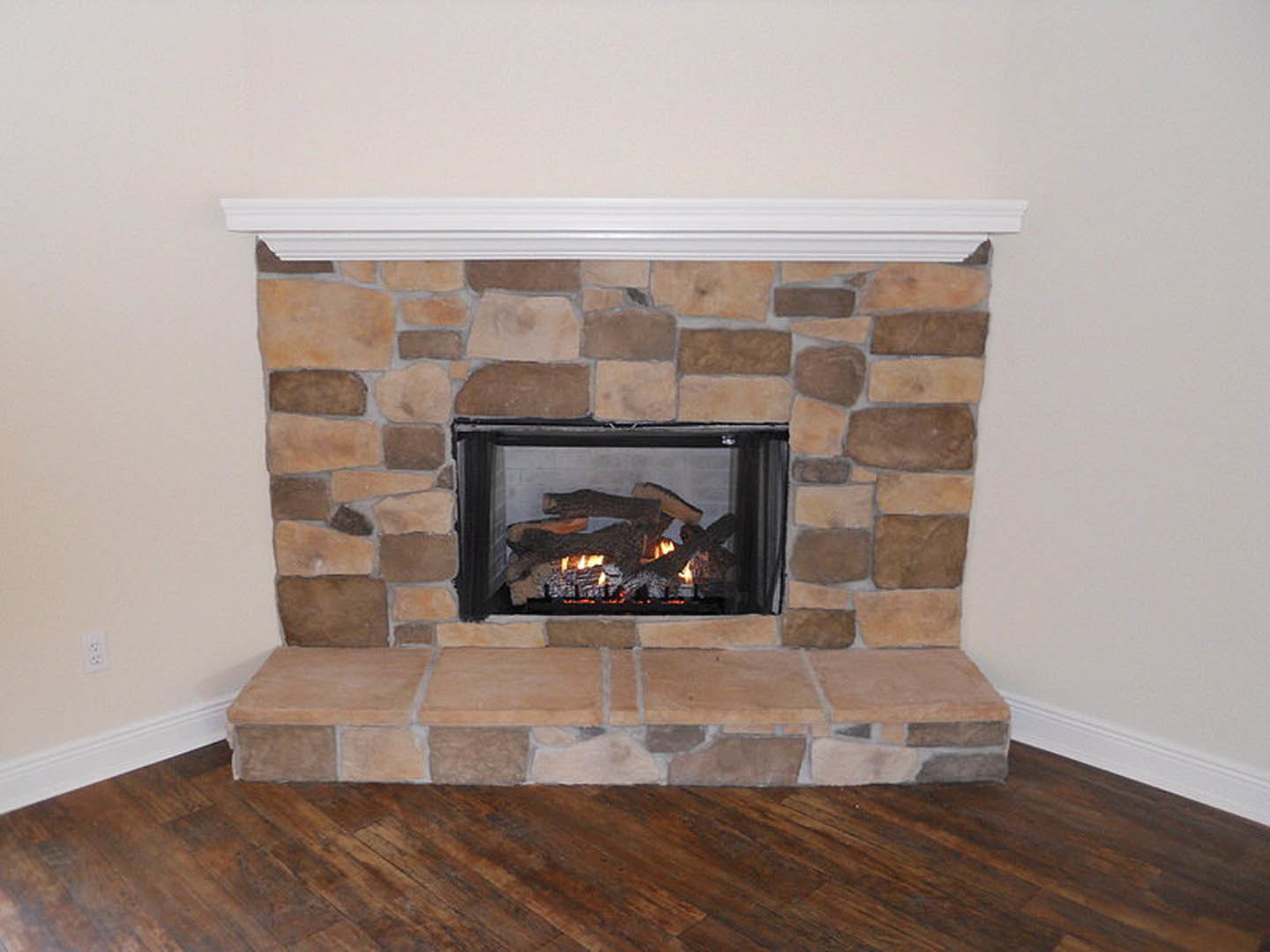 Stone fireplace with burning firewood, wood floor, white walls, and hearth shelf in a cozy indoor living space
