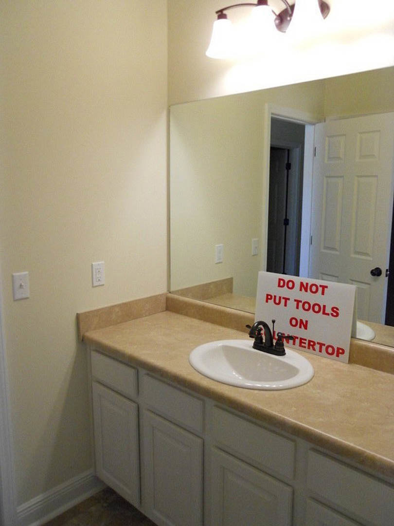 Bathroom with light tile walls, white countertop, chrome faucet, rectangular sink, and small sign placed beside the basin