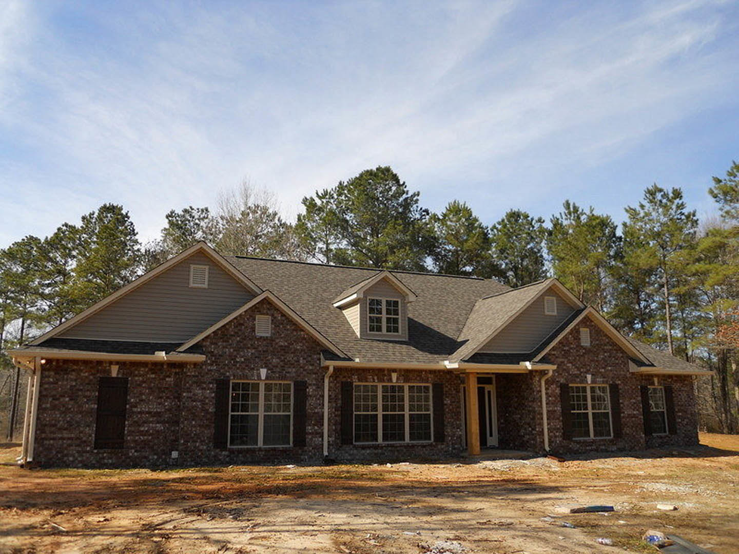 Partially built house with white-framed multi-pane window, exposed wooden framing, unfinished dirt ground, scattered construction debris, and mature trees in the background
