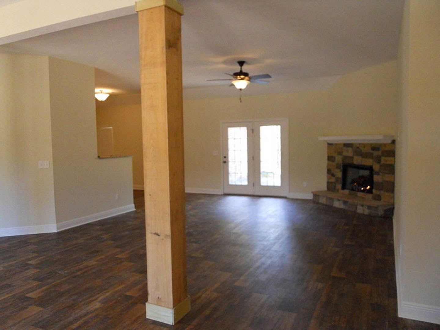 Living room with hardwood flooring, white double doors with glass panes, wooden pillar, plaster walls, ceiling fan with light, and fireplace with burning fire