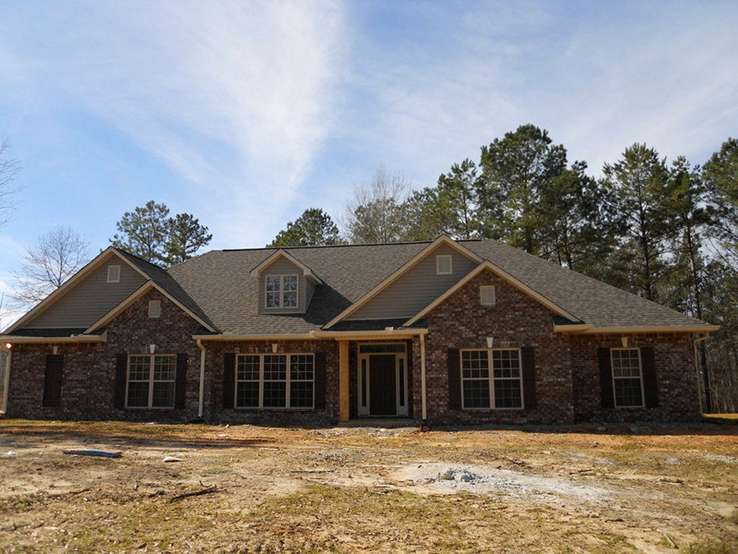 Partially built house with brown roof, multi-pane windows, exposed wooden framing, and dirt construction site, surrounded by leafy trees under cloudy sky