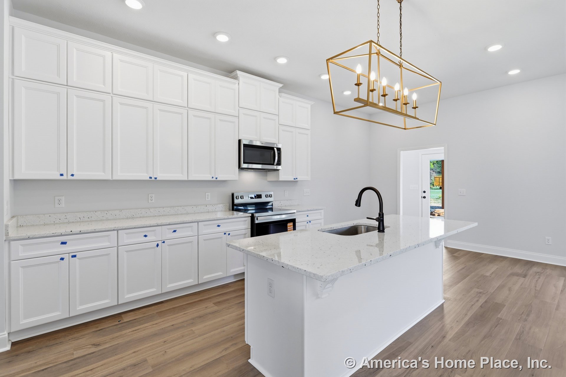 White kitchen with shaker cabinets, black sink and faucet, white island, stainless steel stove and microwave, pendant chandelier, light wood flooring.
