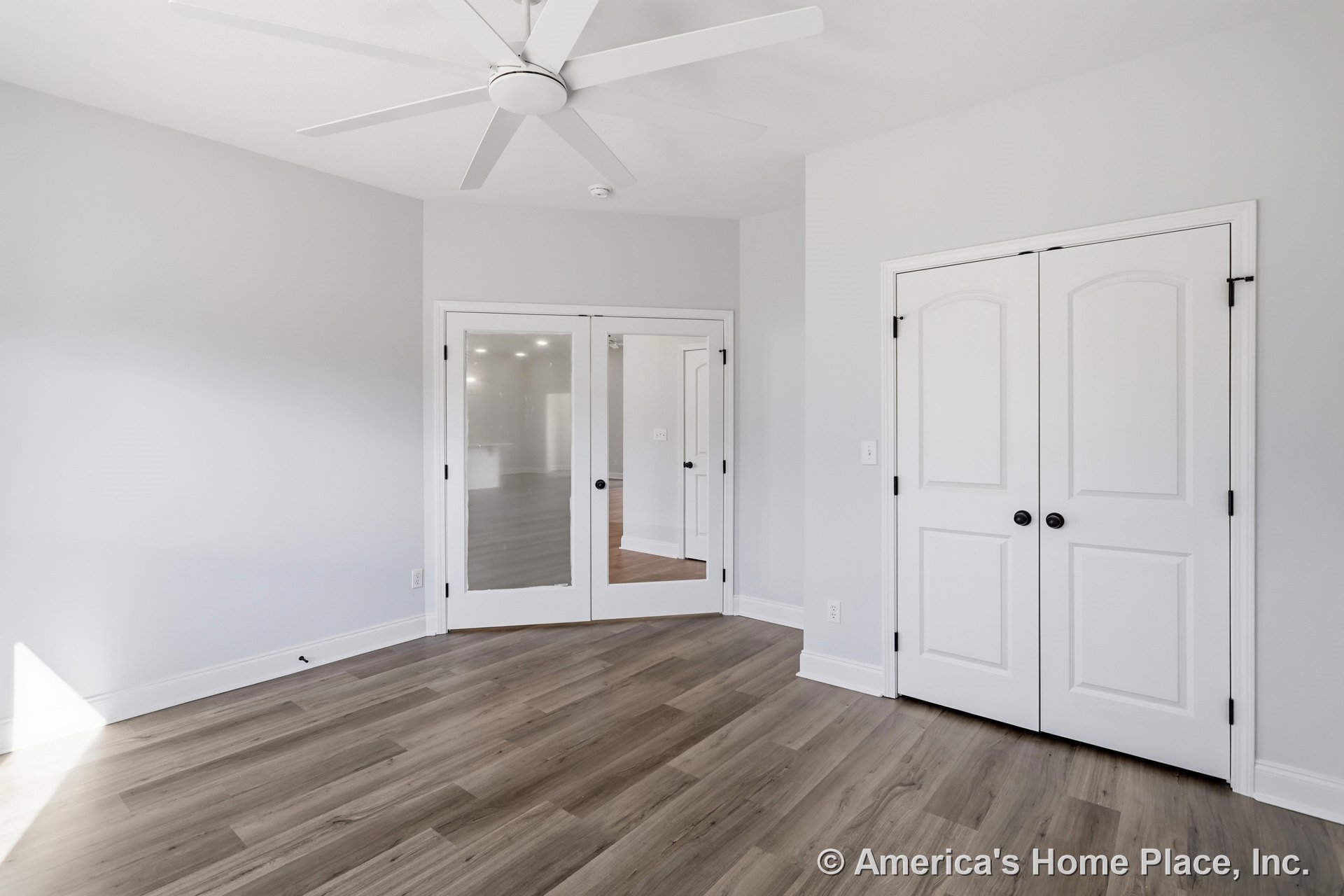 White-walled room with wood flooring, white trim, ceiling fan, fire alarm, window, and white double doors featuring glass panels and black knobs