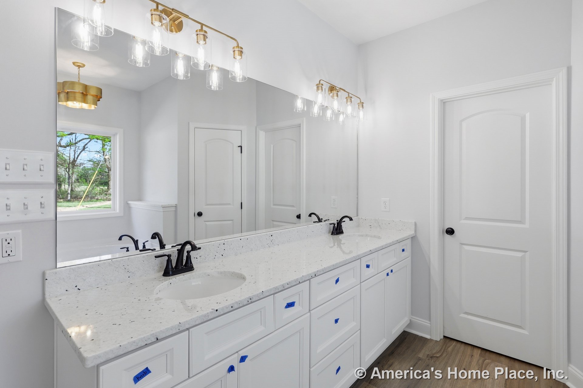 Bathroom featuring white cabinets and white countertops, black faucet and handles, white door with black knob, wall outlet, blue tape with black writing, window with mop placed in