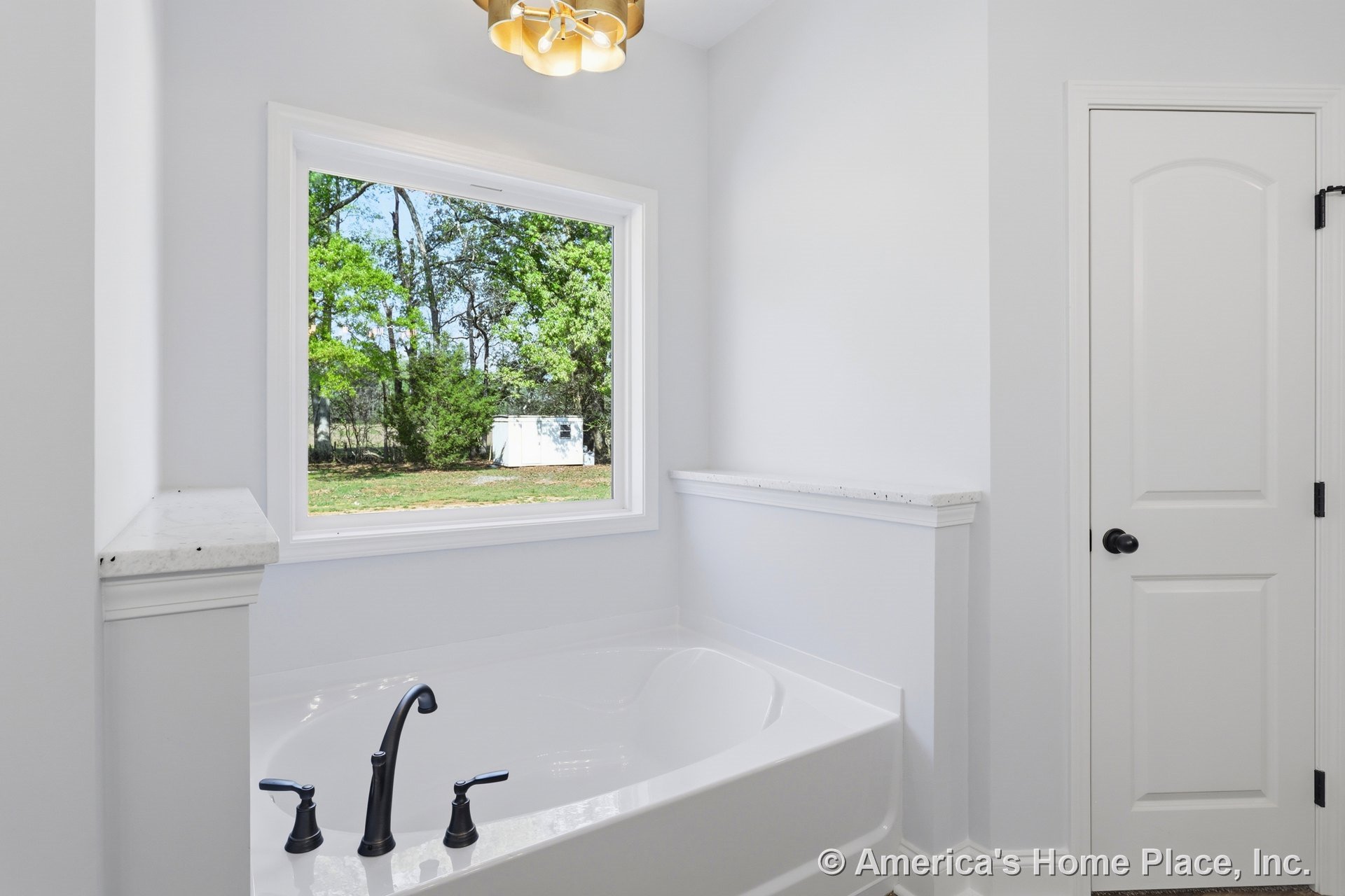 Freestanding white bathtub beneath modern light fixture, positioned near window overlooking trees and small shed, adjacent to white door with black knob in bright bathroom.