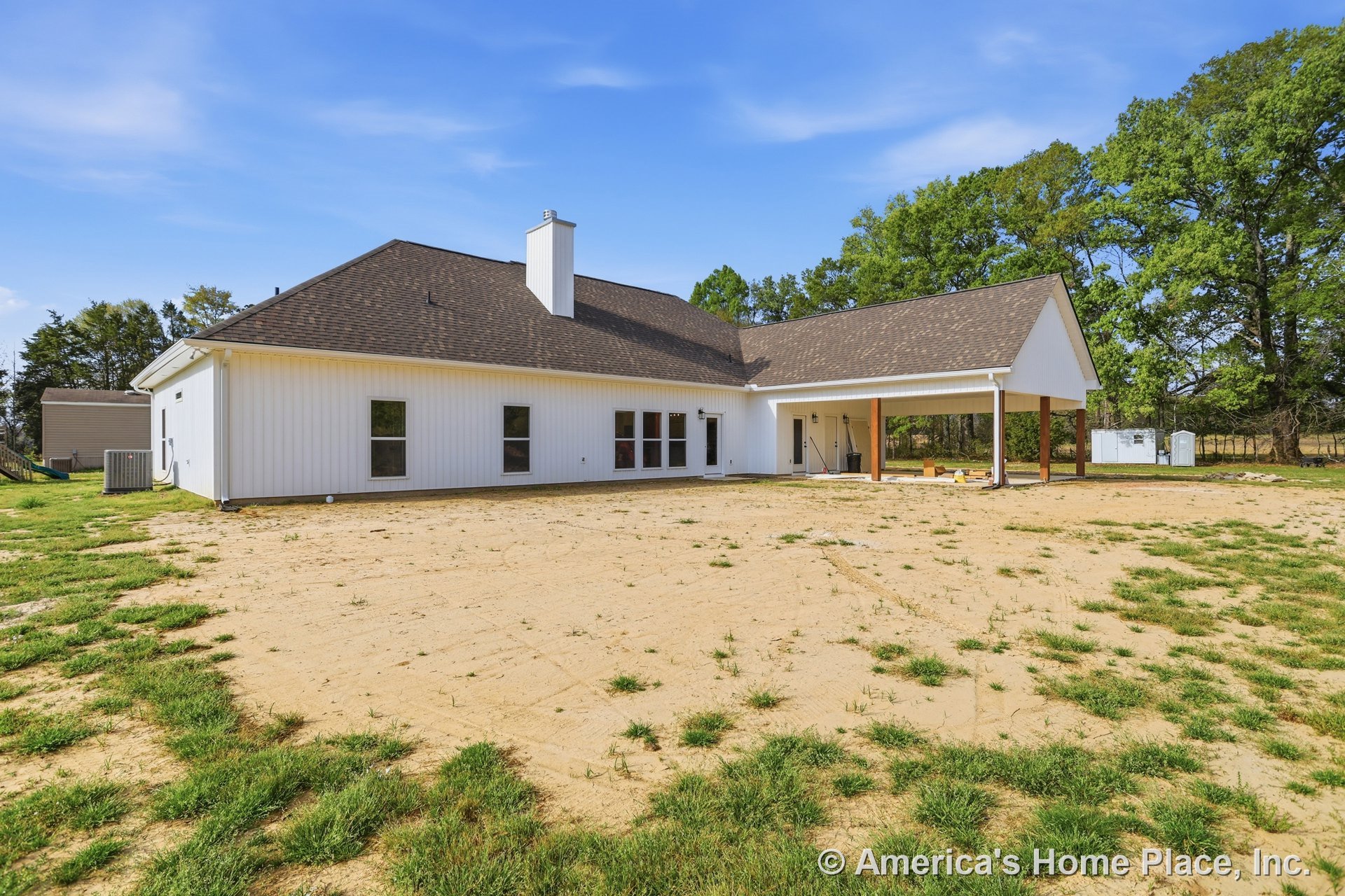 White house with chimney and white-framed windows, surrounded by a dirt yard with patches of grass, metal object in foreground, cloudy sky overhead.