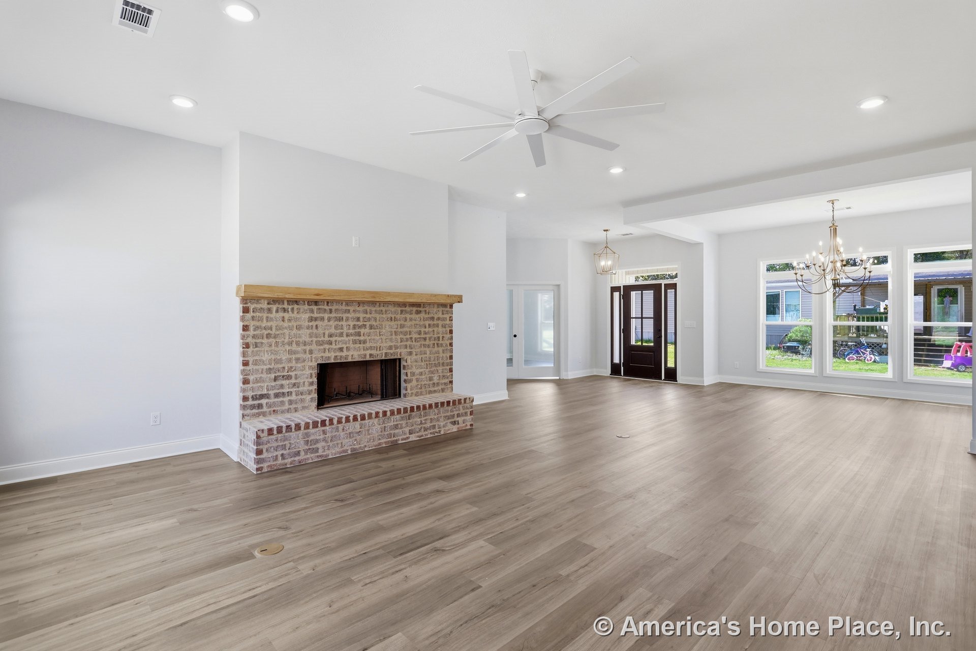 Spacious living room featuring hardwood floors, white ceiling fan with light, central fireplace with white mantel, white walls, door with glass panels, ceiling vent