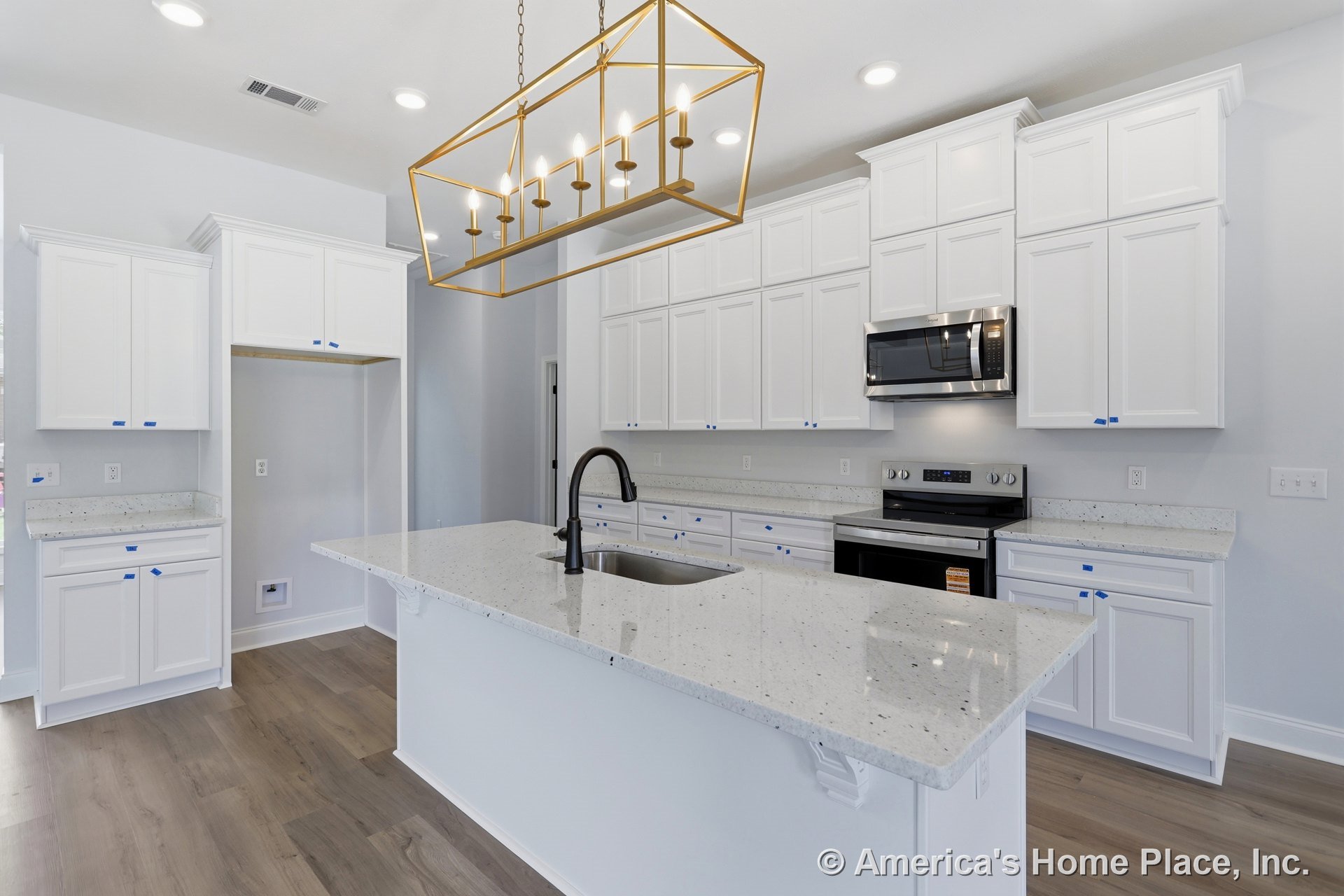 White kitchen with shaker cabinets, large central island featuring a black sink, gold chandelier overhead, stainless steel microwave and stove, light stone countertops, hardwood