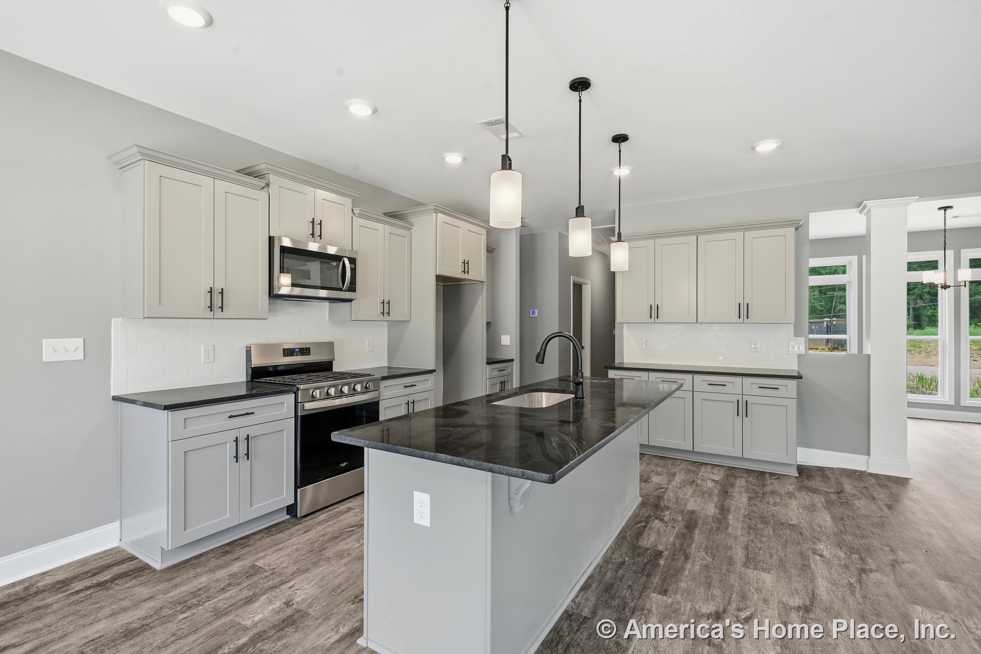 Gray shaker cabinets, black stone countertops on a spacious island, pendant lights overhead, stainless steel appliances, white subway tile backsplash, wood-look flooring, large
