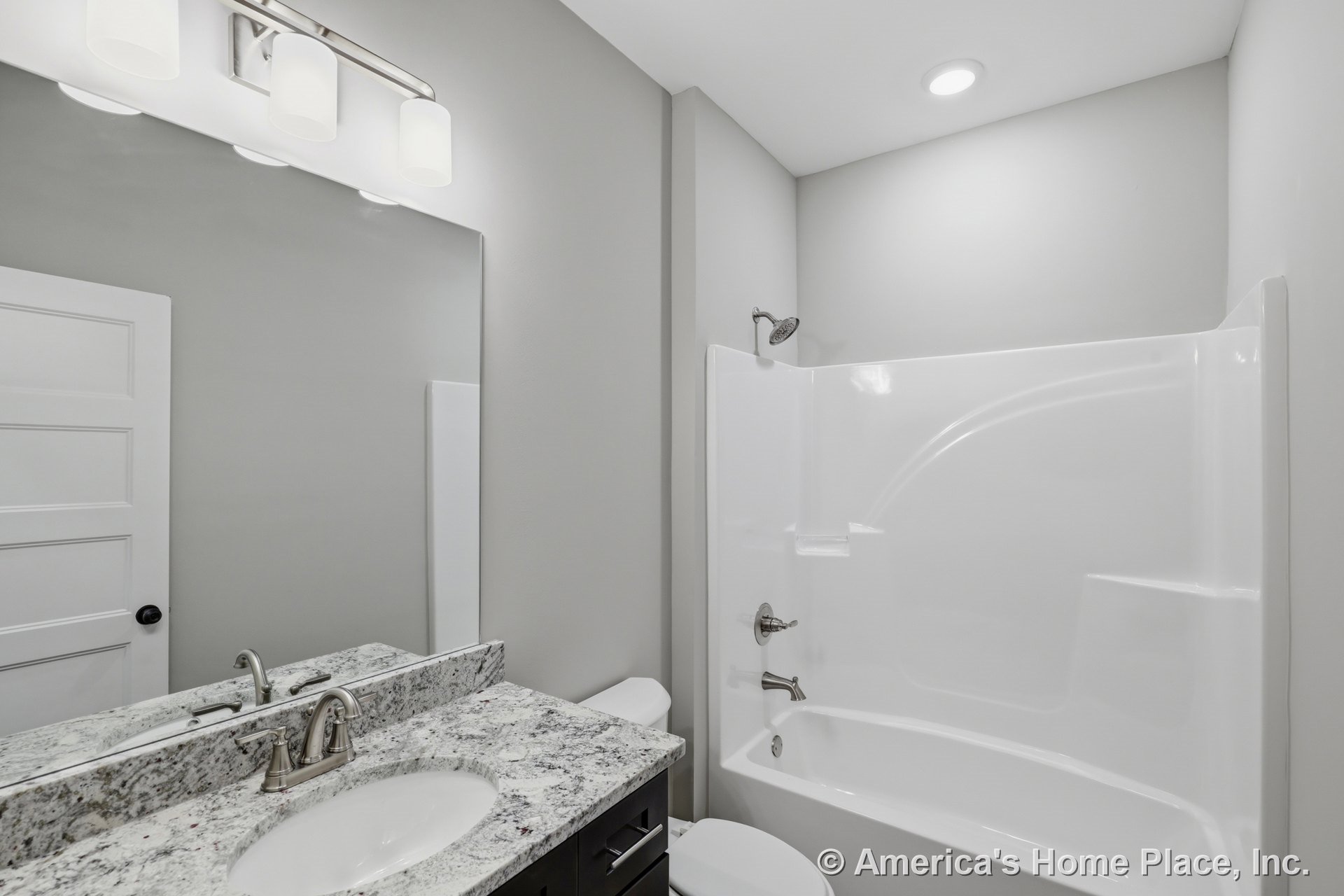 Granite countertop vanity with modern faucet, built-in fiberglass tub and shower combination, large wall mirror above three-light vanity fixture, recessed ceiling light, white