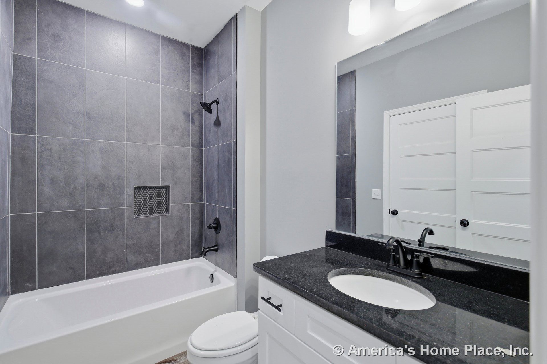 Dark large format tile shower surround with built-in niche, black granite vanity countertop, white shaker cabinetry, matte black plumbing fixtures, paneled interior door