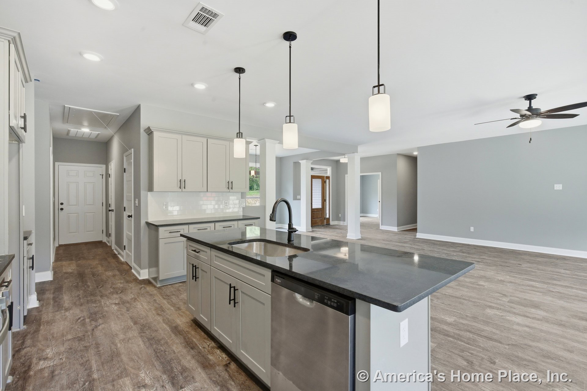 Shaker-style kitchen cabinets with dark stone countertops and pendant lights above an island sink, opening to a spacious living area with wood-look flooring, ceiling fan, large