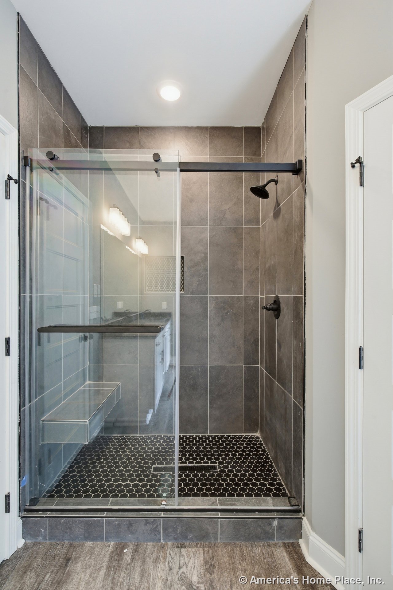 Walk-in shower with dark hexagonal floor tile, large format tile walls, built-in bench, glass sliding door, matte black fixtures, and recessed ceiling lighting.