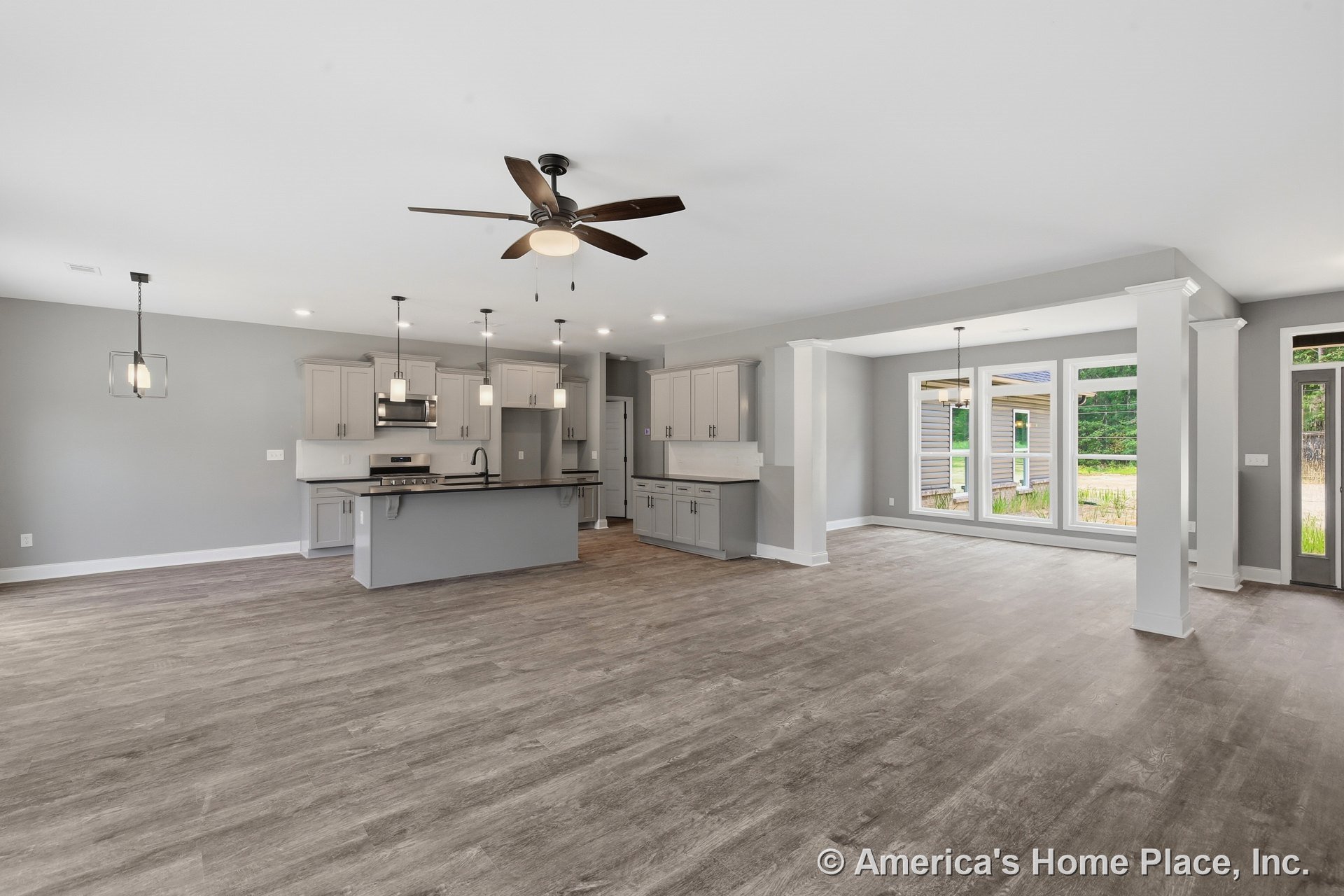 Open-concept kitchen and living area with gray shaker cabinets, pendant lights above the island, wide plank wood-look flooring, large picture windows, ceiling fan with light