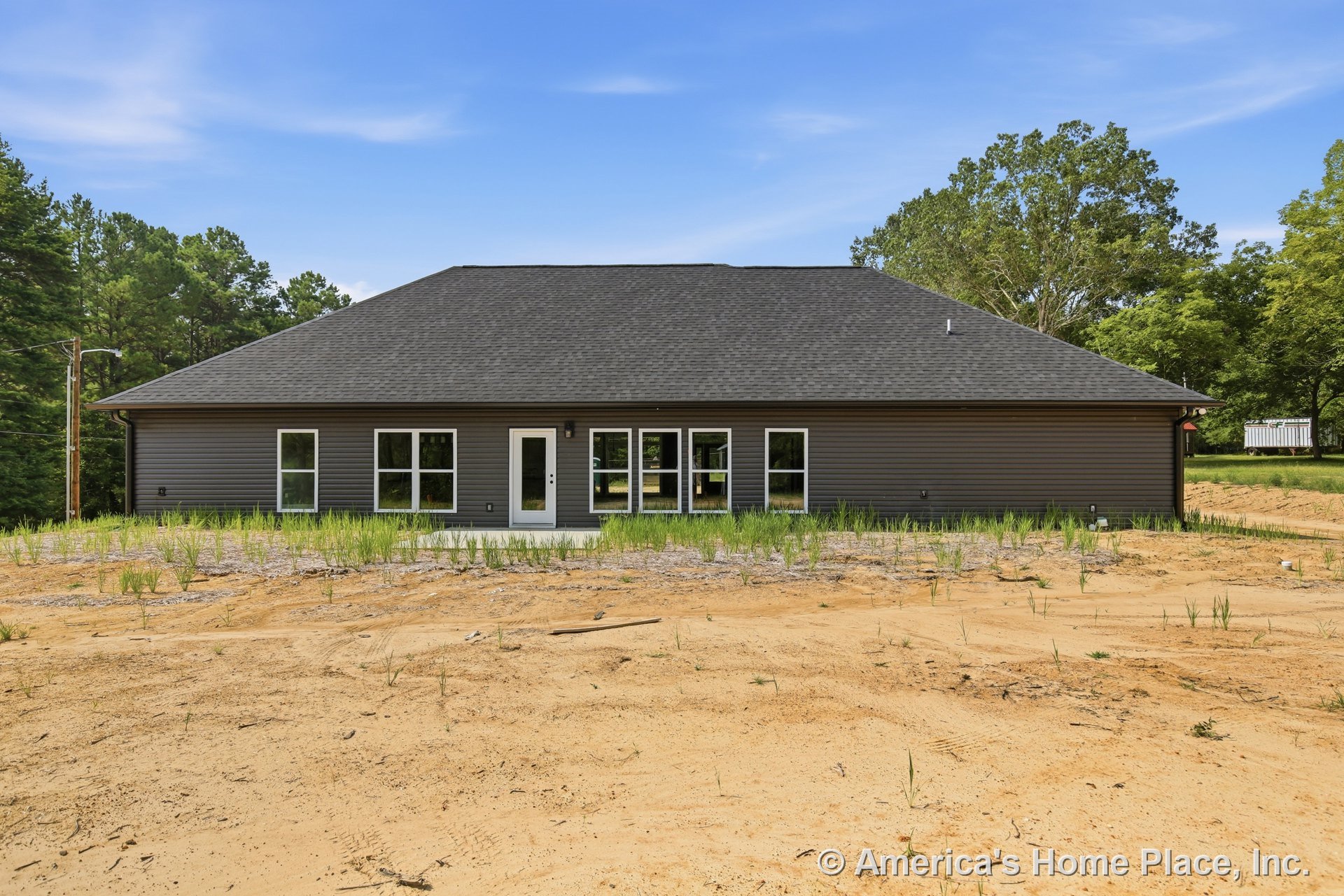 Rear exterior with dark horizontal siding, shingled hip roof, multiple large windows, single exterior door, concrete patio, and unfinished yard.