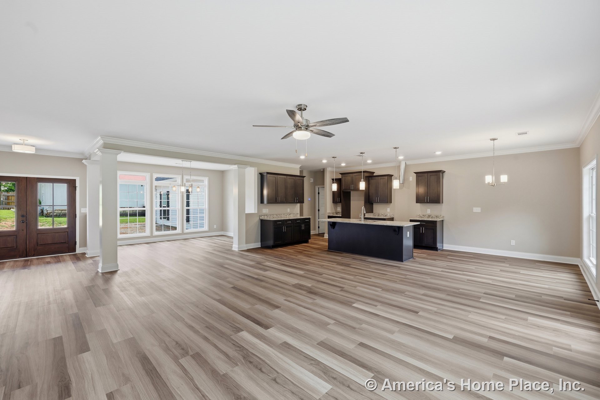 Open-concept living and kitchen area with dark wood cabinets, pendant lights above a central island, expansive window wall with trim, light wood plank flooring, double entry doors