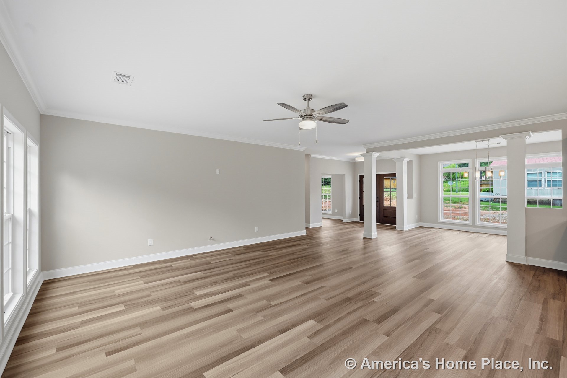 Open concept living area with light wood plank flooring, expansive double windows, ceiling fan with light, white crown molding, entry columns, neutral walls, and recessed lighting.