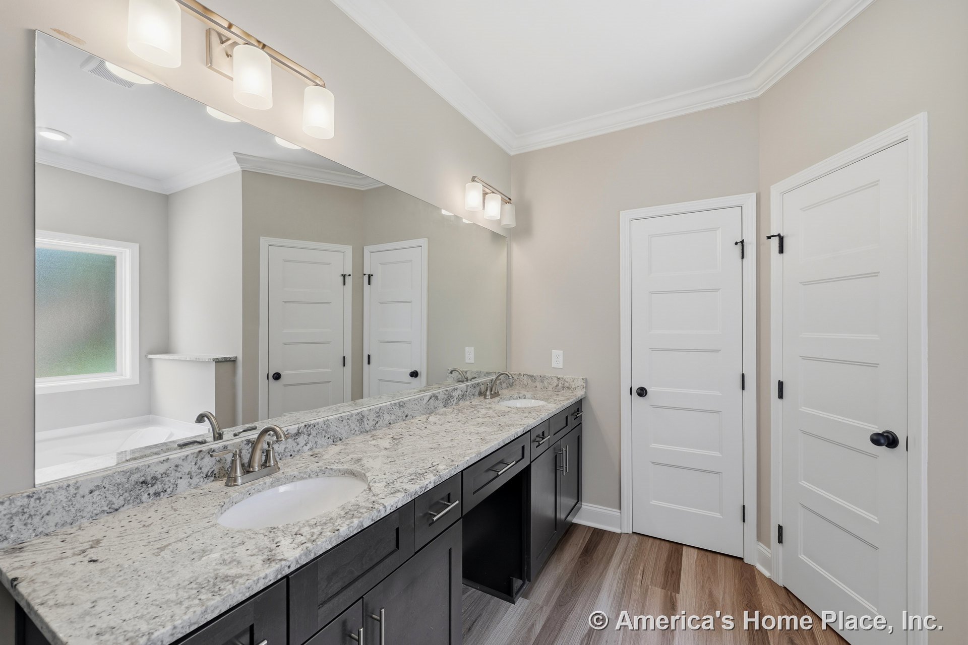 Double vanity with granite countertop and dark cabinetry, dual undermount sinks with modern brushed nickel faucets, large wall mirror, frosted window, white paneled doors