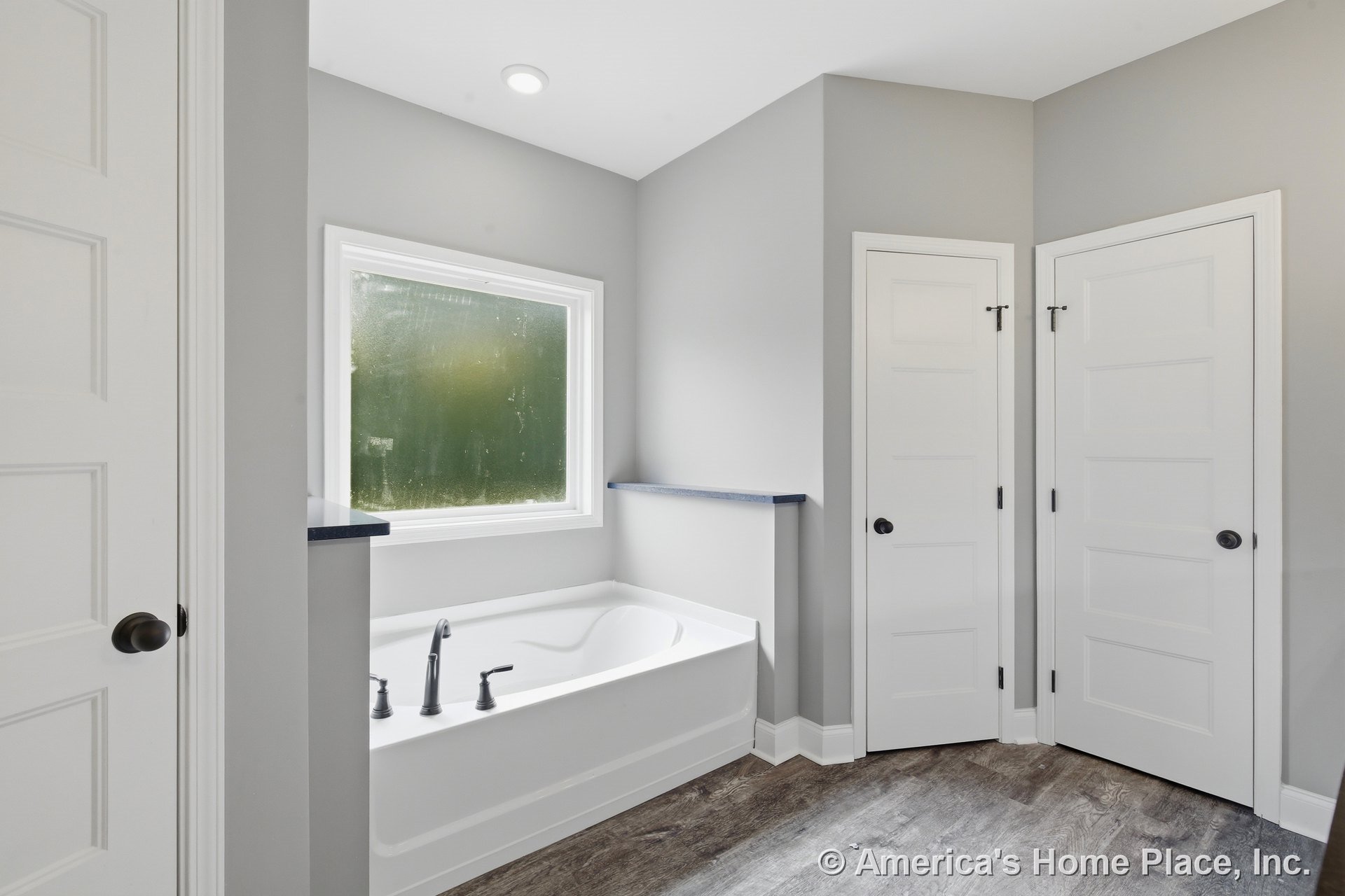 Built-in soaking tub with brushed nickel fixtures set beneath a large frosted window, three paneled interior doors, gray painted walls, wood-look vinyl plank flooring, recessed