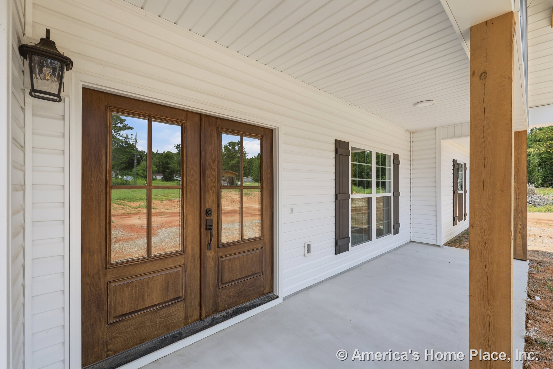 Double wooden entry door framed by white vinyl siding, large windows with dark shutters, covered porch ceiling with lighting fixture, concrete porch floor, and wooden support post.