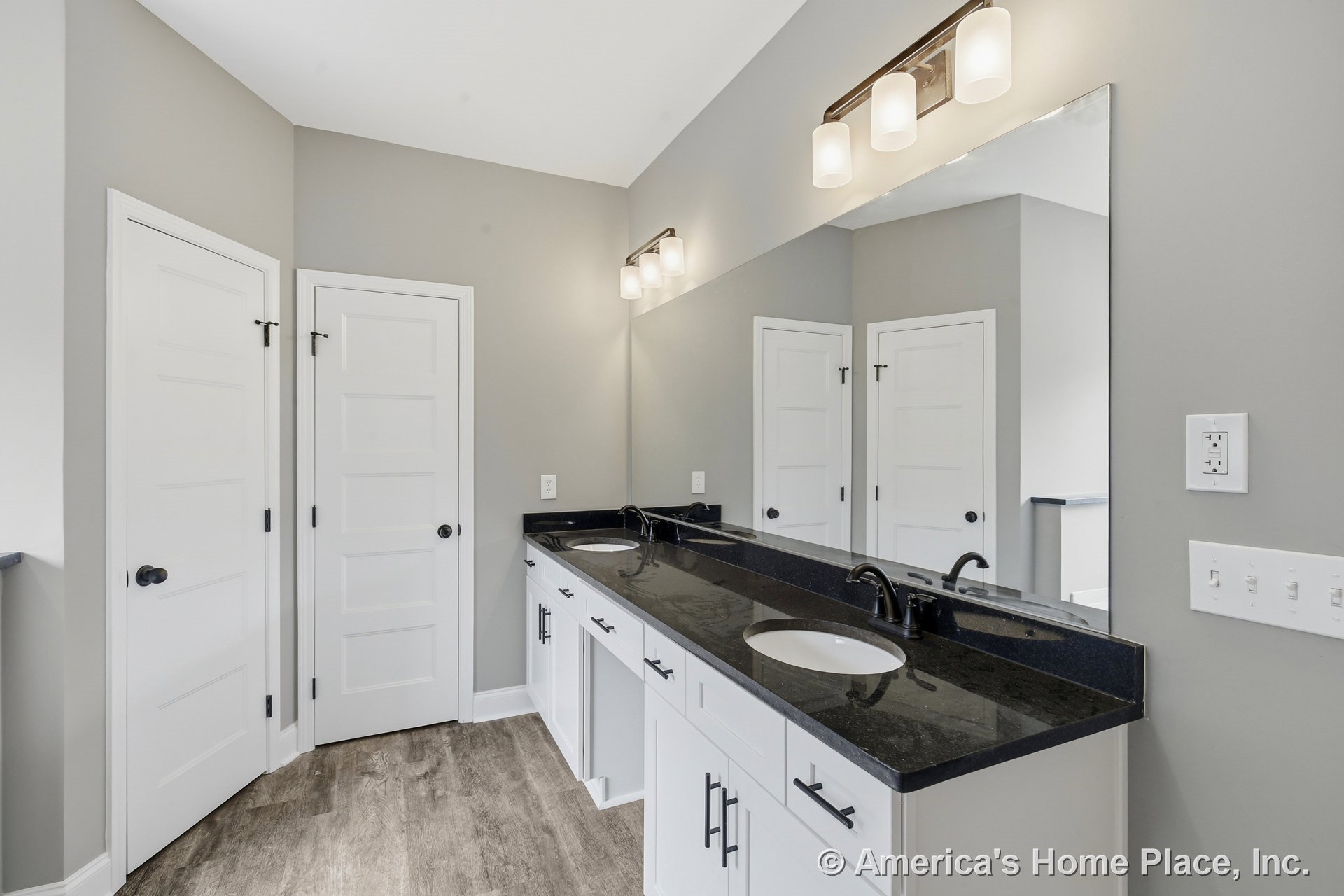 Double vanity with black stone countertop, white shaker cabinets, undermount sinks, matte black hardware, large wall mirror, modern vanity lighting, wood-look flooring, panel
