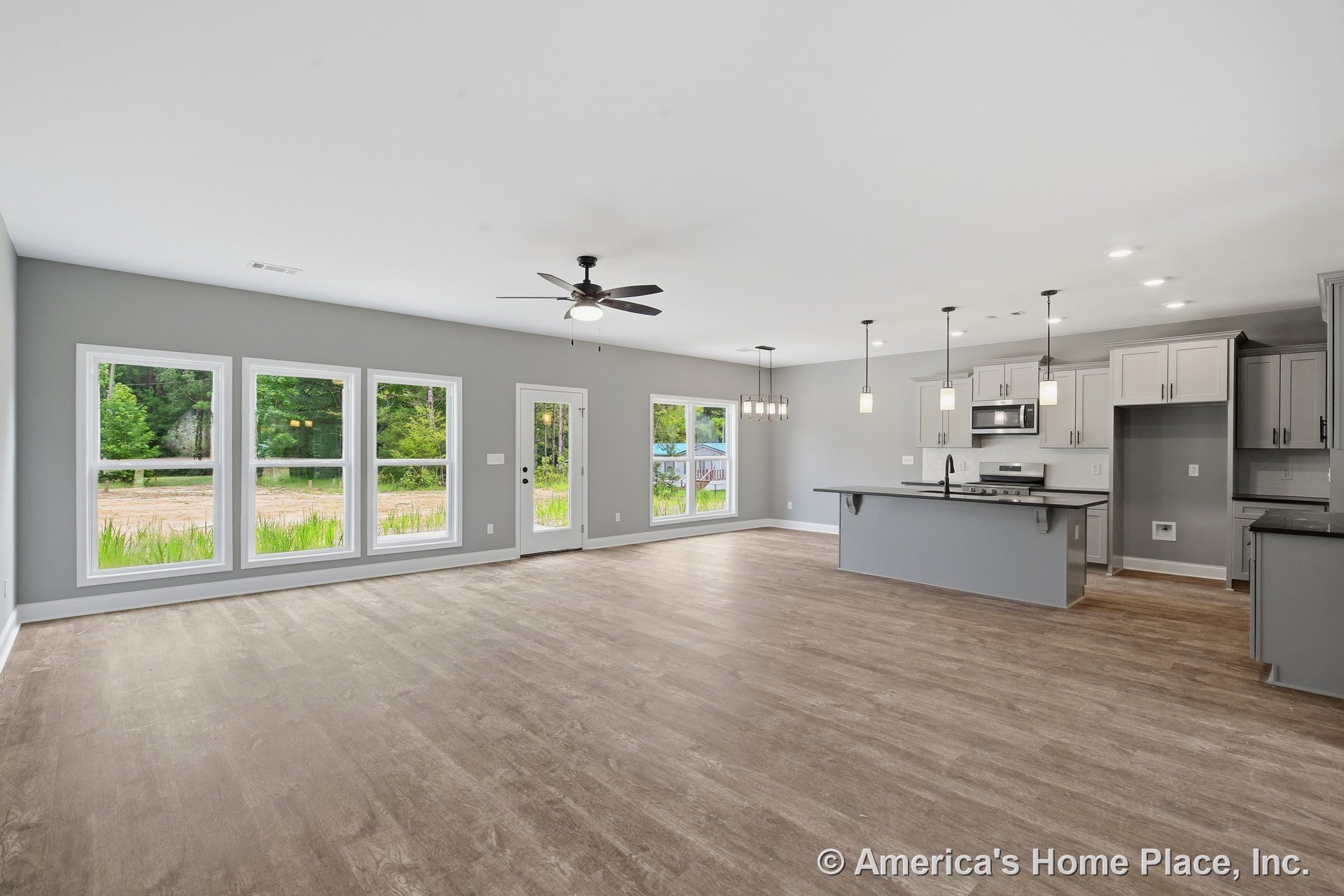 Open-concept living area with four-panel picture windows, gray shaker kitchen cabinets, pendant and recessed lighting, wood-look vinyl plank flooring, kitchen island with overhang