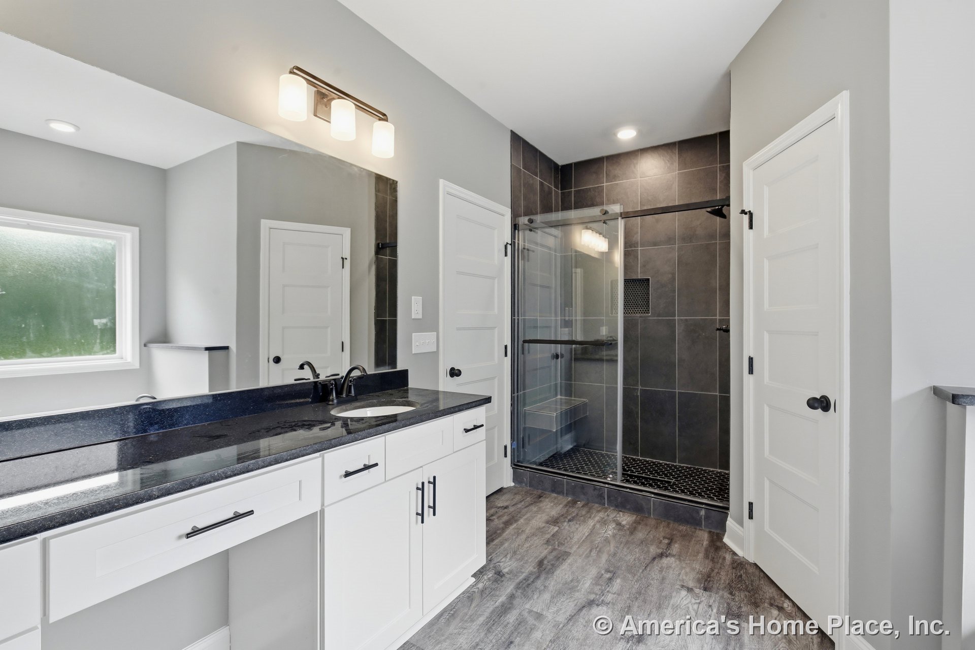 Walk-in glass shower with dark tile walls, double-sink vanity featuring black stone countertops and white shaker cabinets, wood-look flooring, modern lighting, window, and built-in