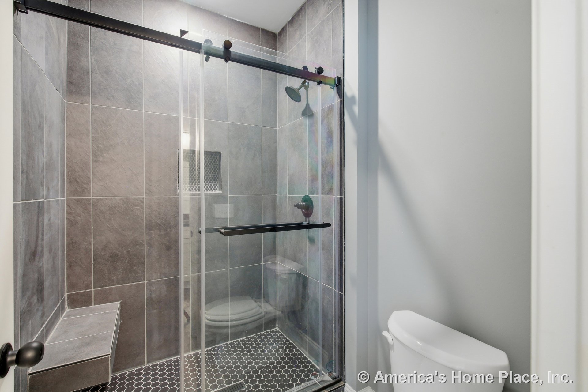 Walk-in shower with large gray tile walls, glass sliding door, built-in bench, hexagonal tile floor, mosaic tile shower niche, and adjacent white toilet.