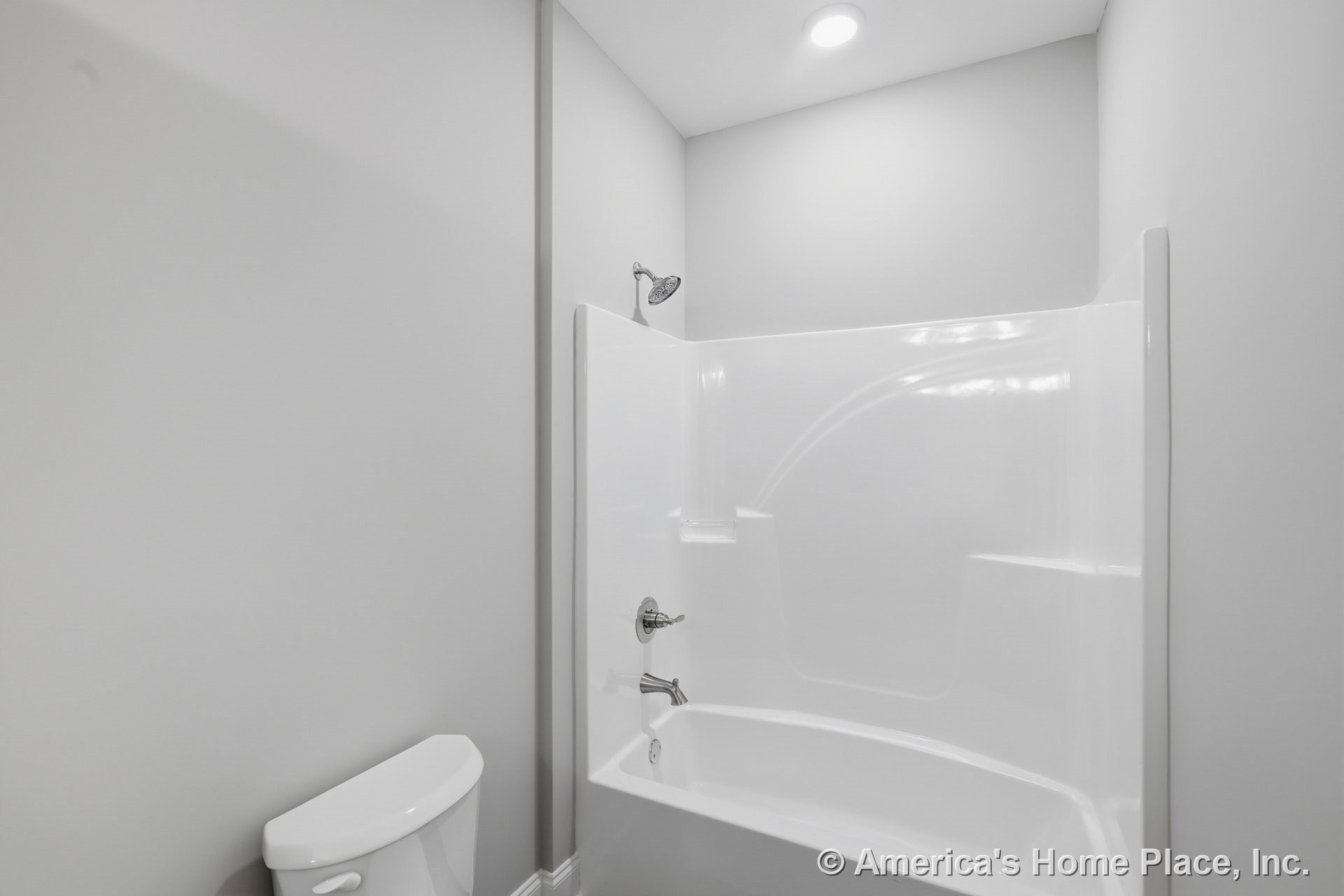 Built-in fiberglass tub and shower combination with chrome showerhead and faucet, recessed ceiling light, white toilet, and light gray walls.