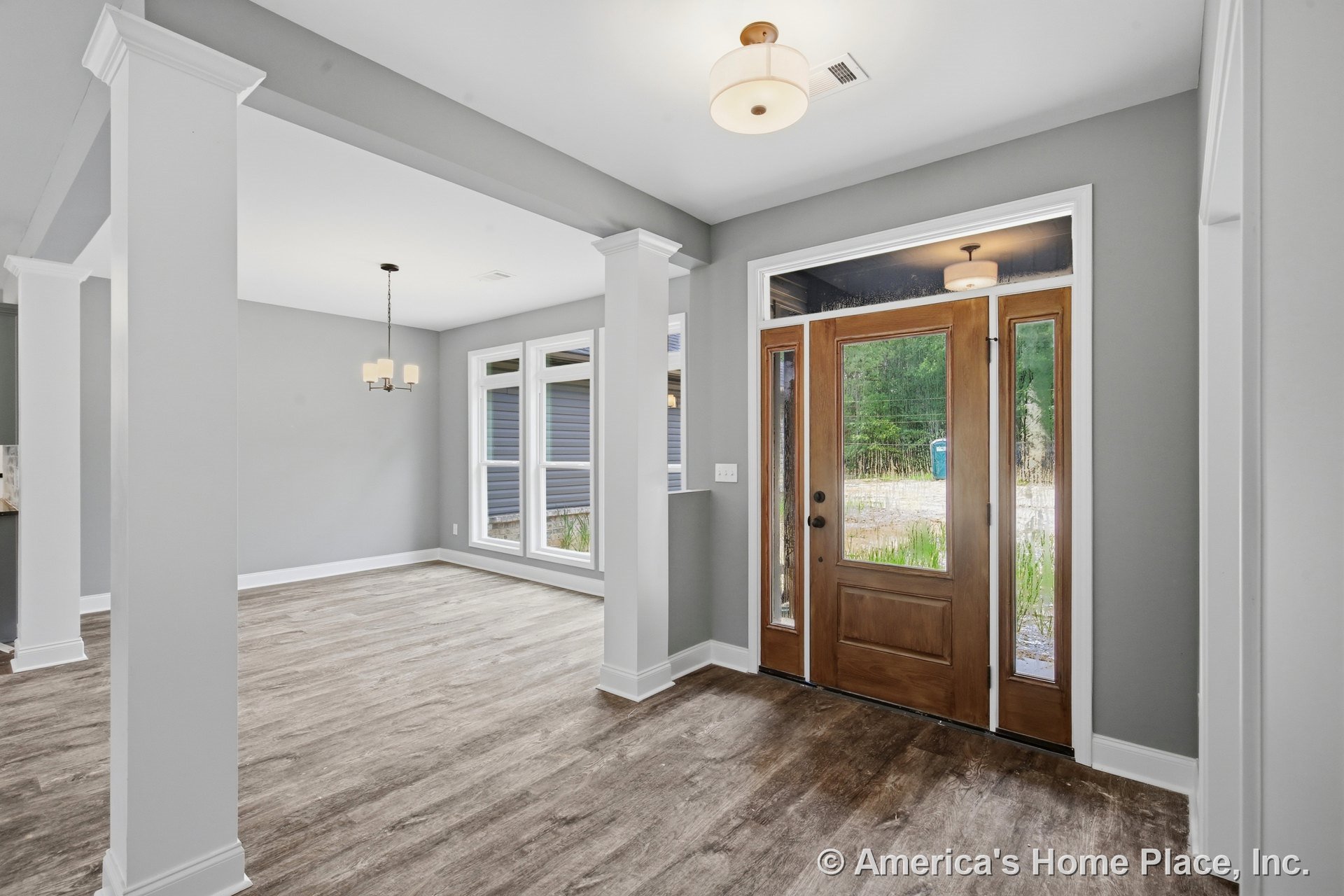 Wooden front door with glass sidelights, gray painted walls, white trim and columns, wood-look vinyl flooring, built-in ceiling lights, large windows in adjacent dining area, open