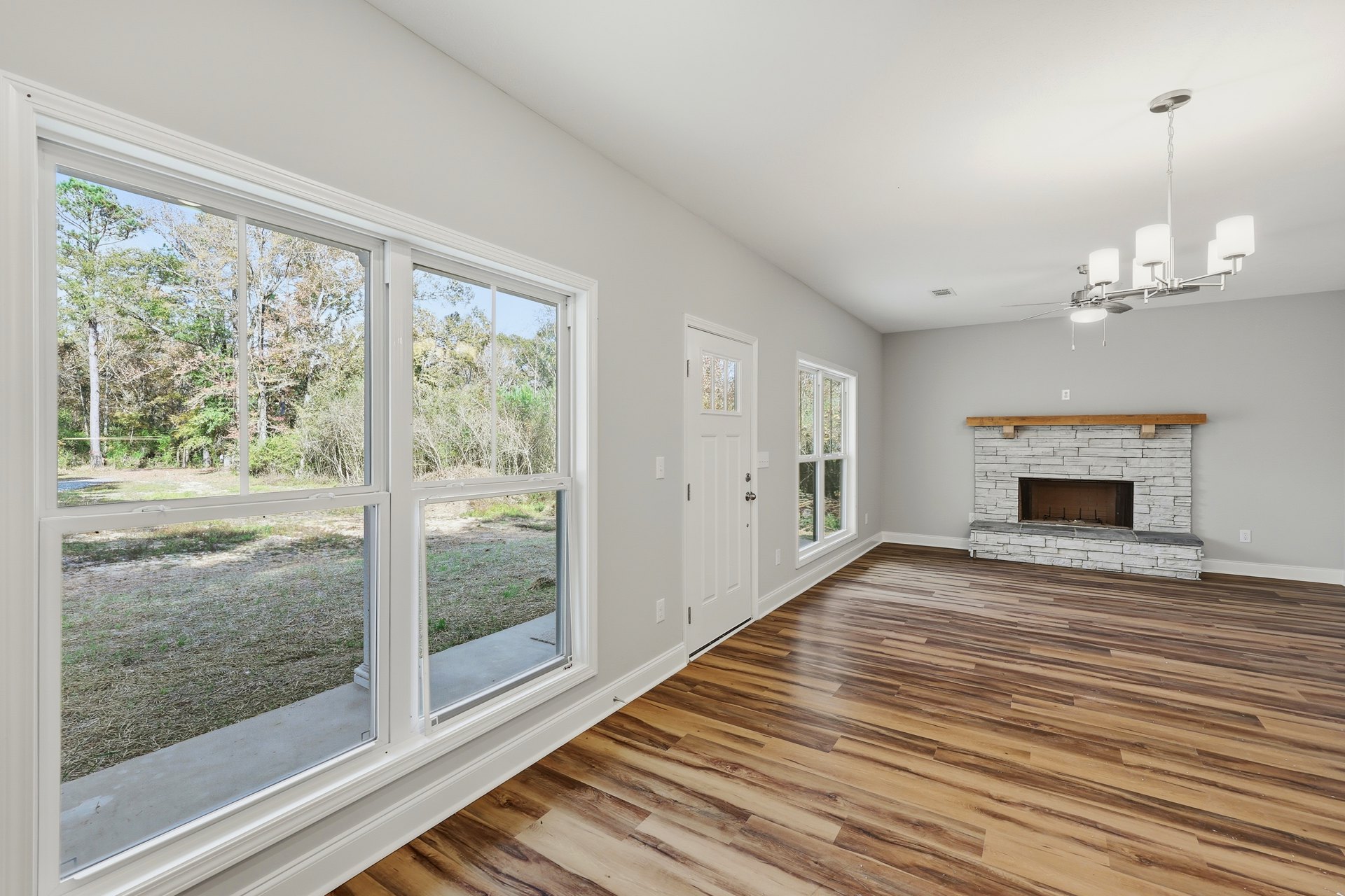 Living room with wood flooring, white walls, a stone fireplace topped by a wood beam mantel, large windows showing trees outside, and a door leading to another space.