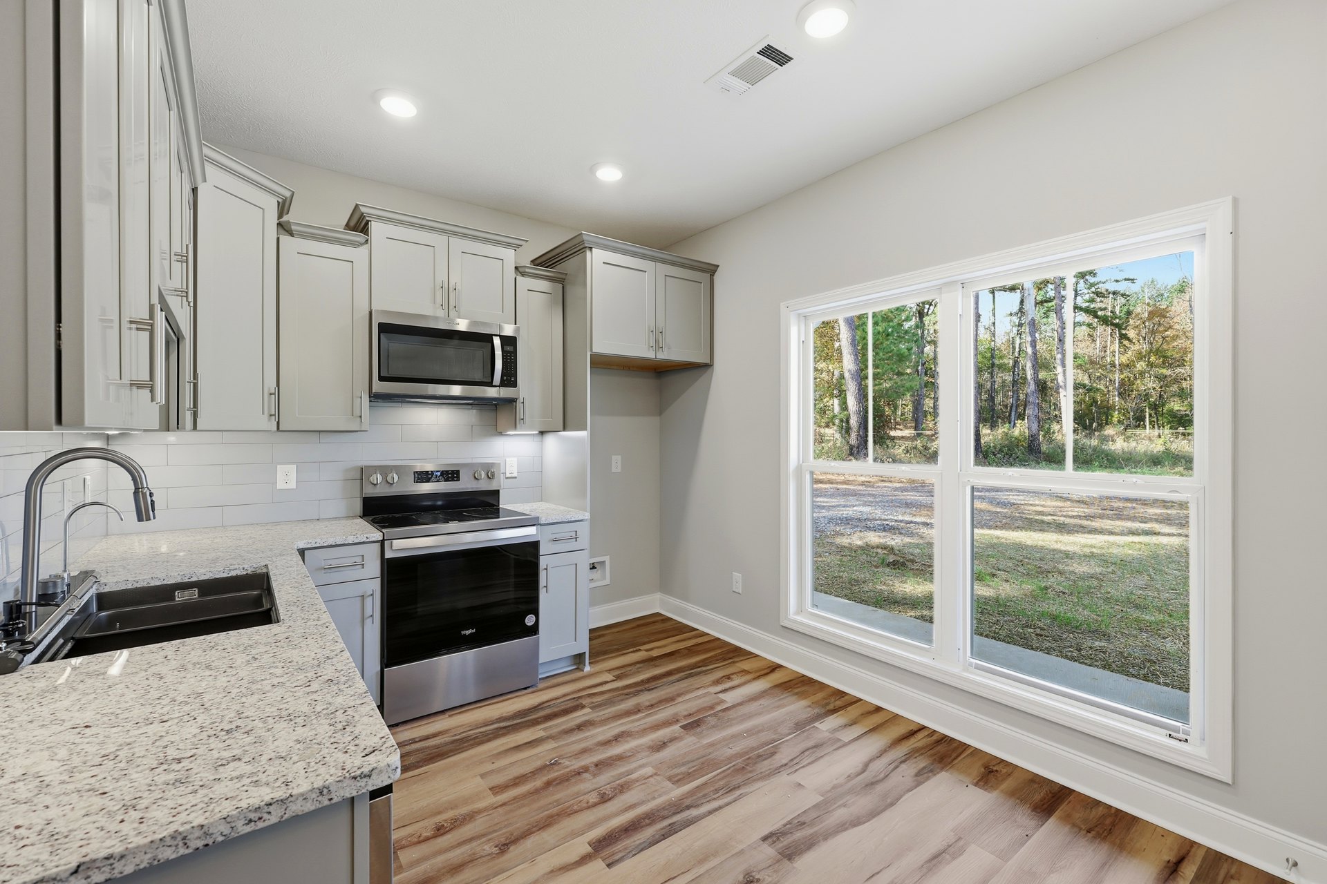 Kitchen with wood flooring, white cabinetry, stainless steel stove and oven, built-in microwave with glass door, large window above sink, chrome faucet, light-colored countertops